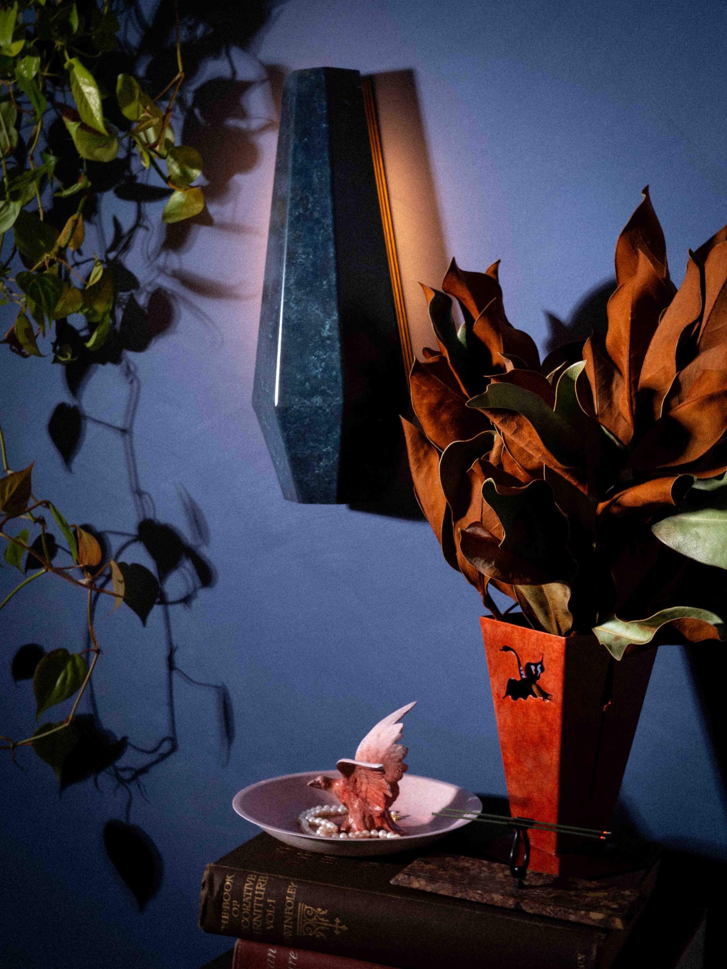 A decorative setup including a pitcher with dried leaves, a couple of books, a small pumpkin, a ceramic pitcher, a black box with an American flag design, and green and brown artificial foliage, set against a blue wall.