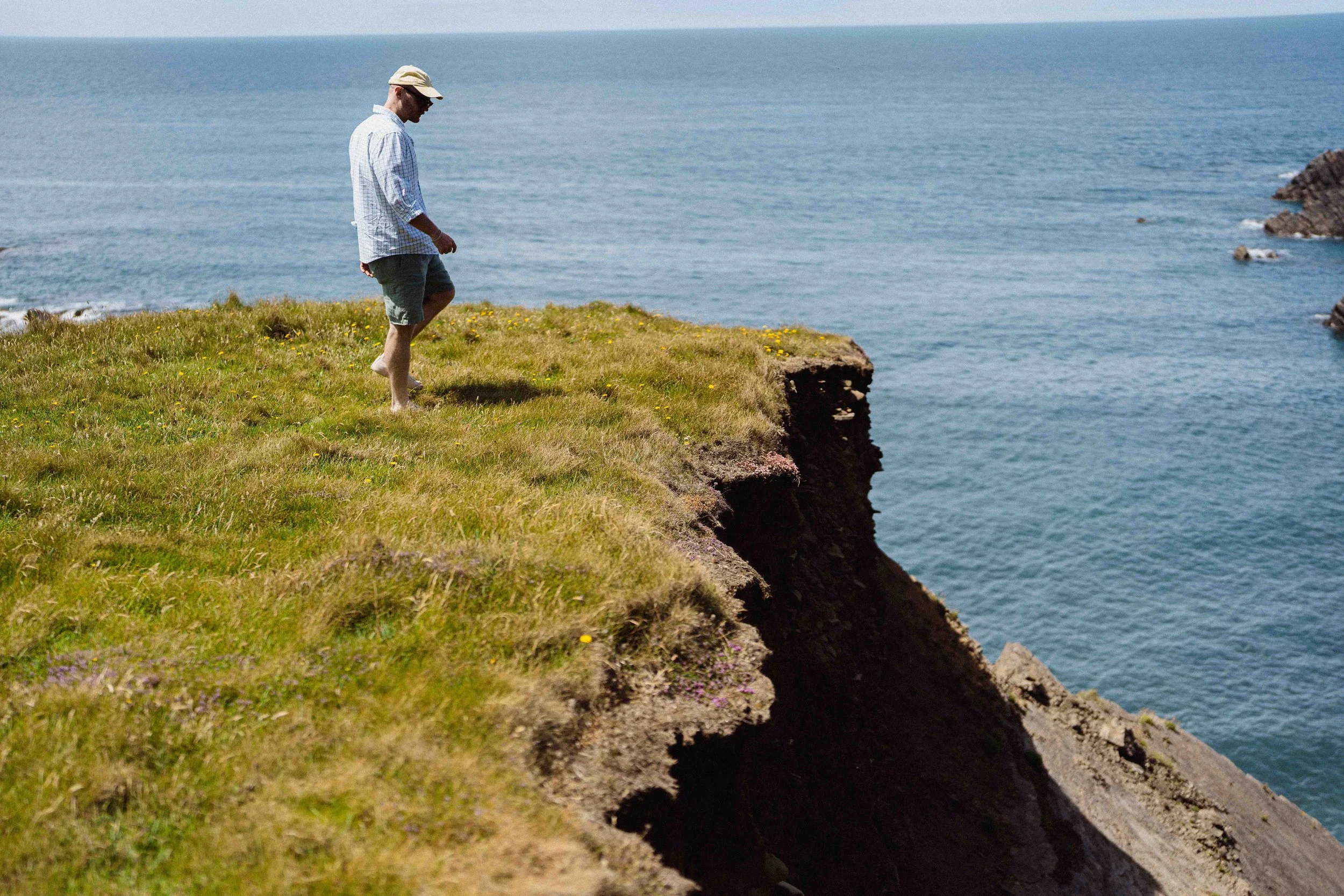 Man walking along the grassy cliff edge overlooking the ocean, wearing a light hat, sunglasses, a checkered shirt, and shorts.