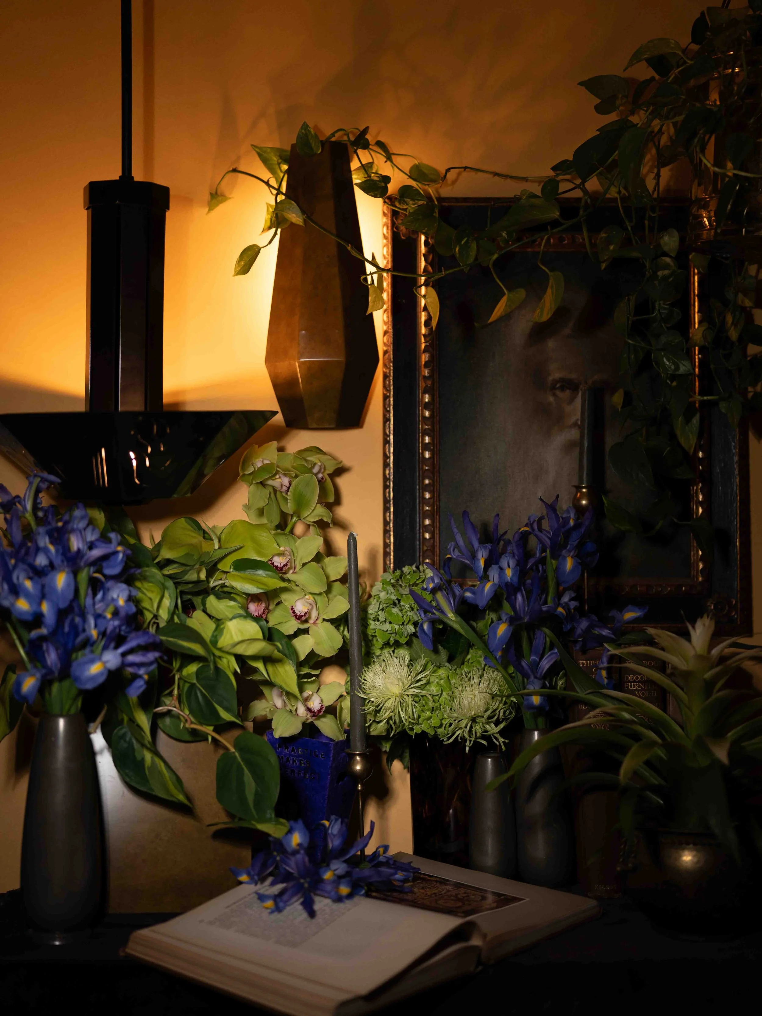 A still life scene showing a variety of flowers in vases, an open book, a mirror, and a black lamp with yellowish lighting creating warm shadows on the wall.