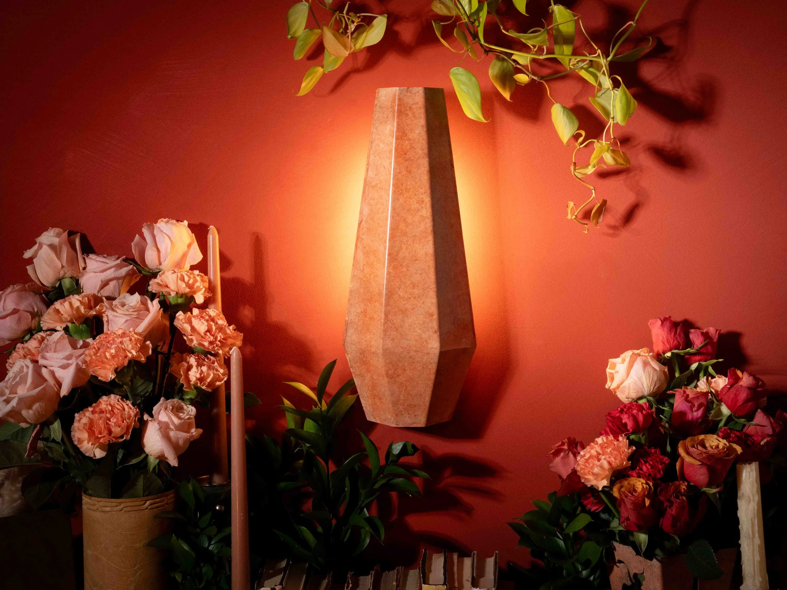 A tall, tapered, marble or stone obelisk sculpture against a deep red wall, surrounded by a variety of pink, peach, and red flowers and green foliage.
