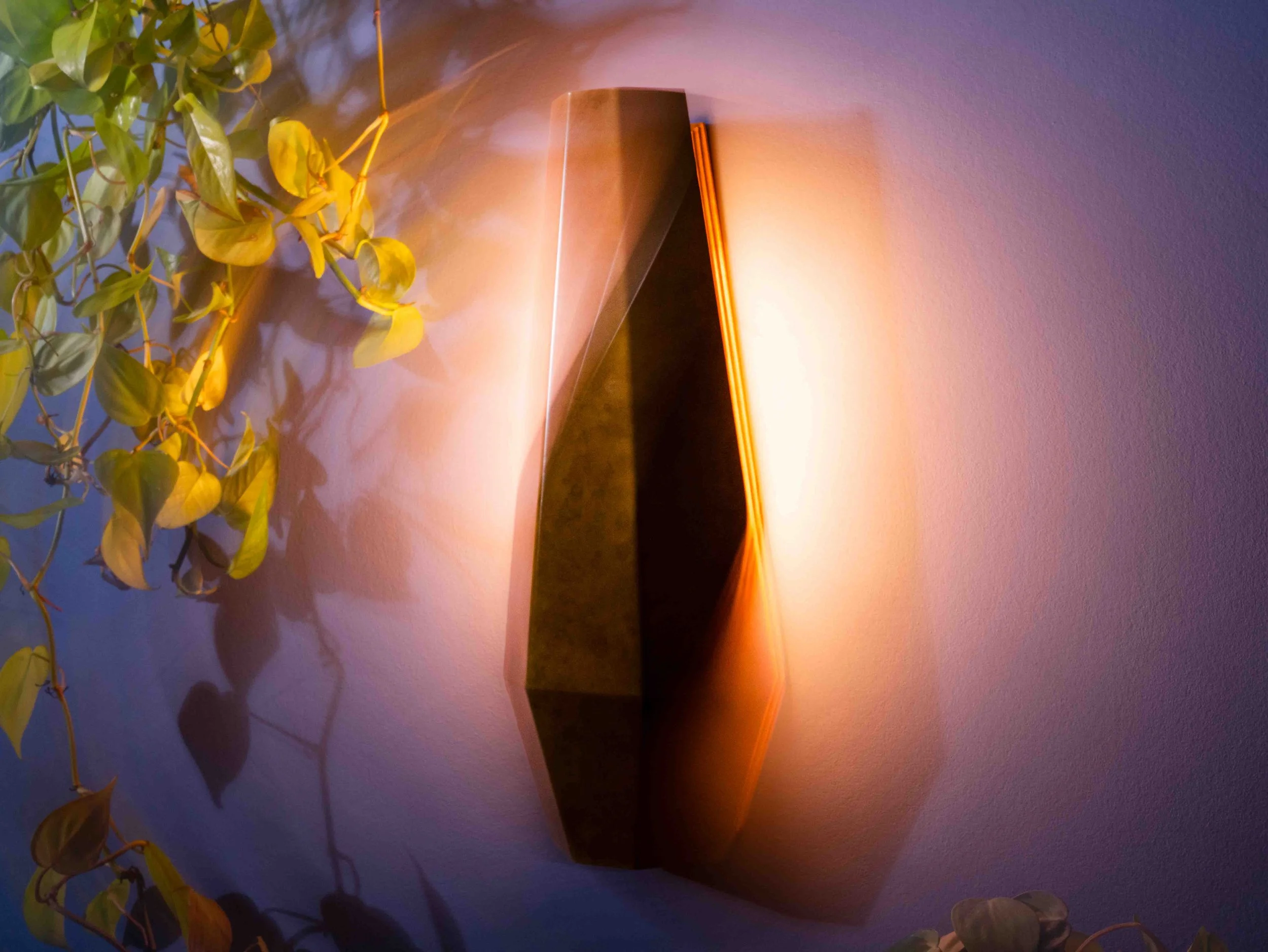 Wall-mounted light fixture with warm orange glow next to a plant with green and yellow leaves on a lavender wall.