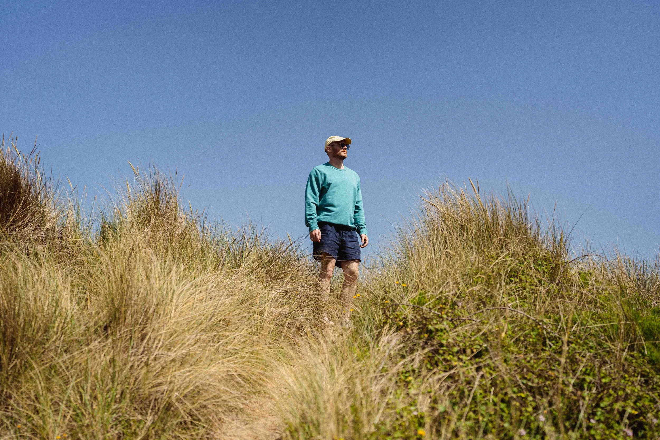 A man wearing sunglasses, a beige cap, a turquoise long-sleeve shirt, and dark shorts standing among tall grasses under a clear blue sky.
