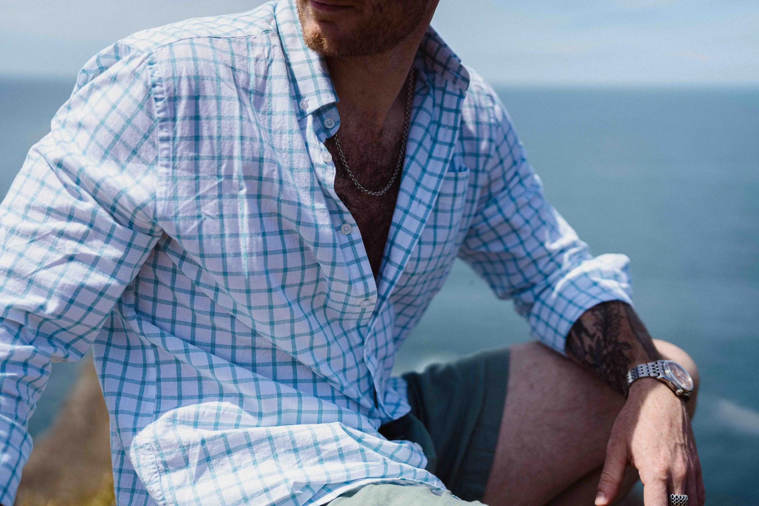 Close-up of a man wearing a blue and white checkered shirt, with a silver chain necklace, sitting outdoors with an ocean background.