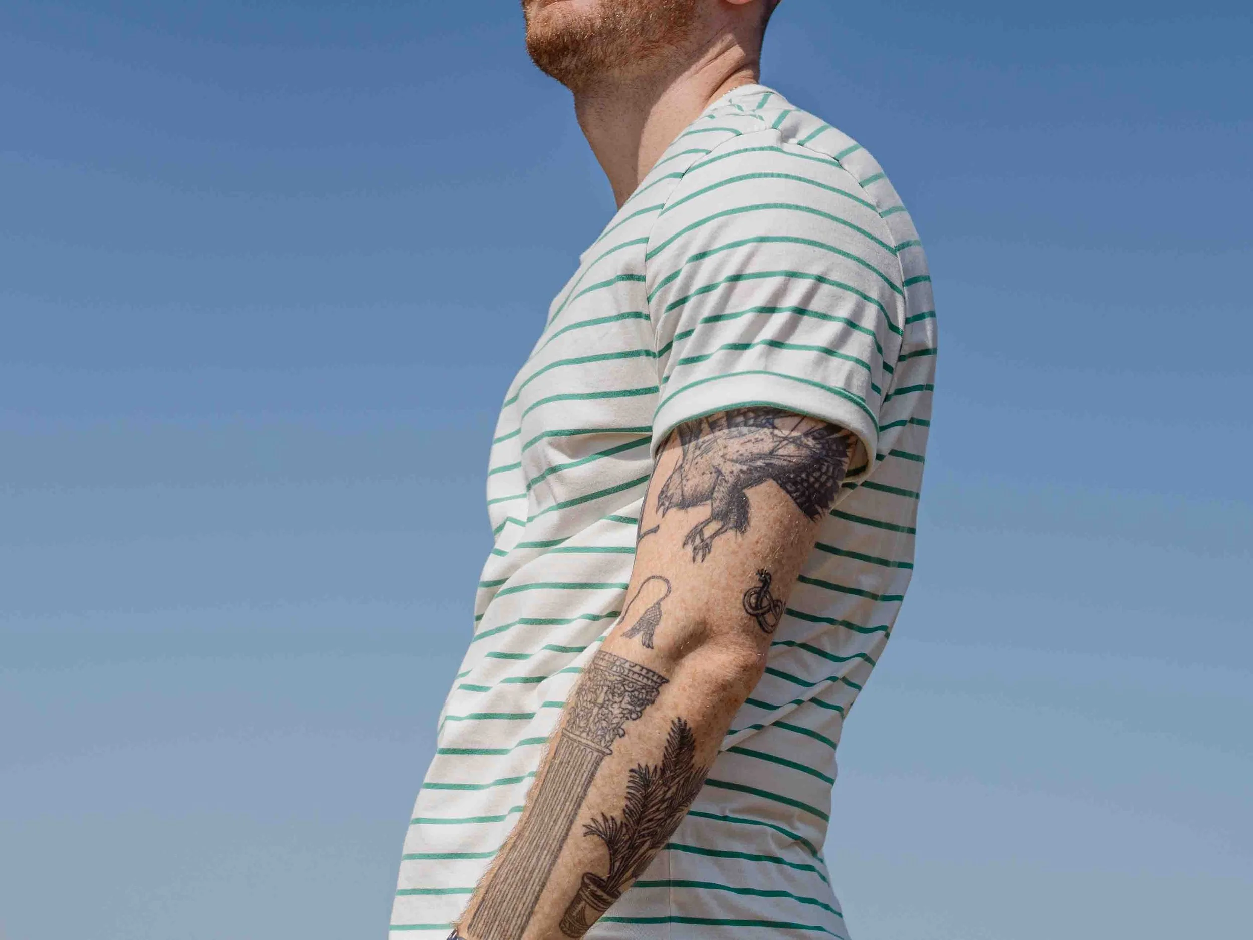 A man with tattooed arms wearing a white t-shirt with green horizontal stripes, standing outdoors against a bright blue sky.