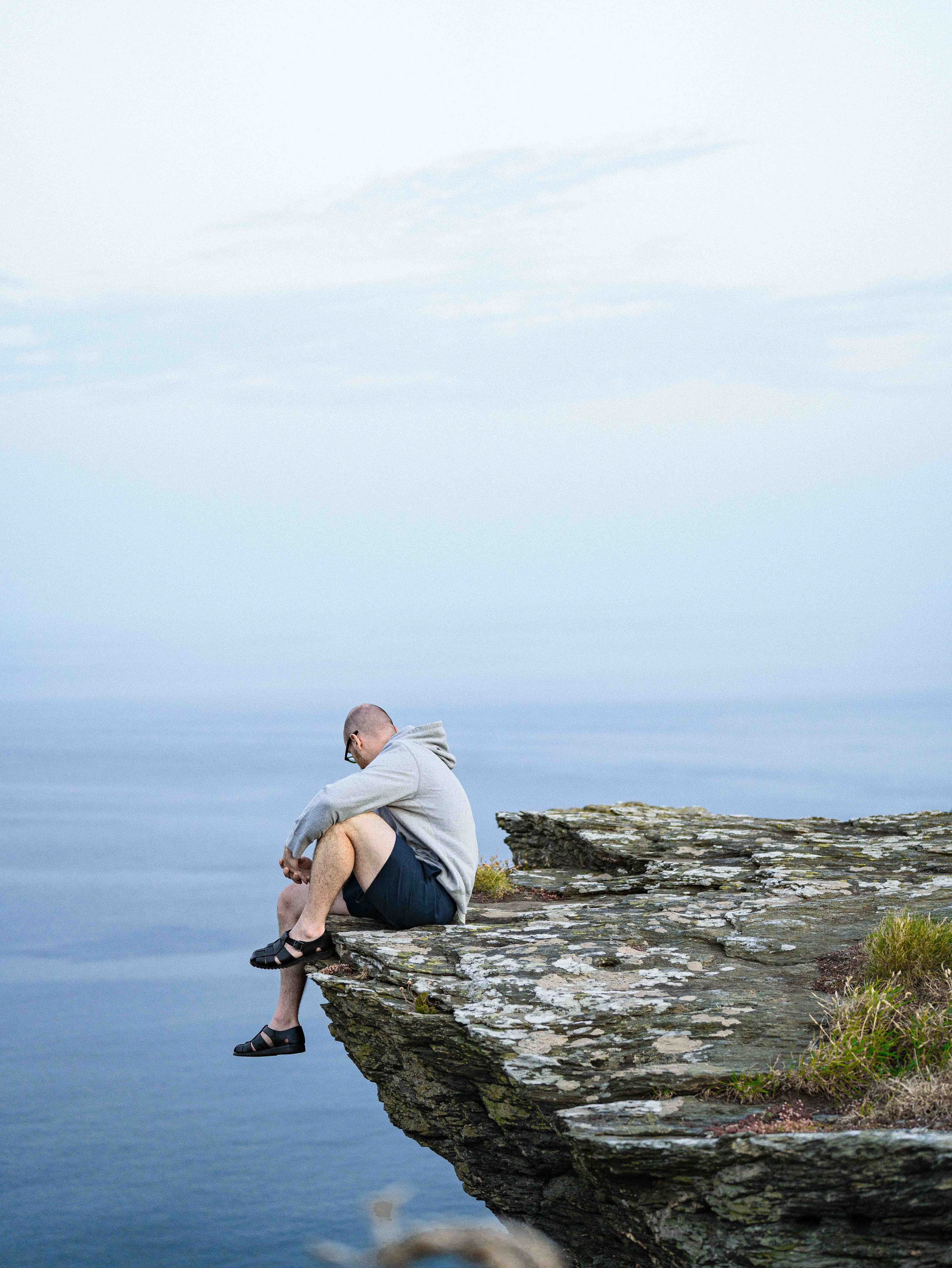 A man sitting on the edge of a rocky cliff, overlooking a calm body of water, with a cloudy sky above.