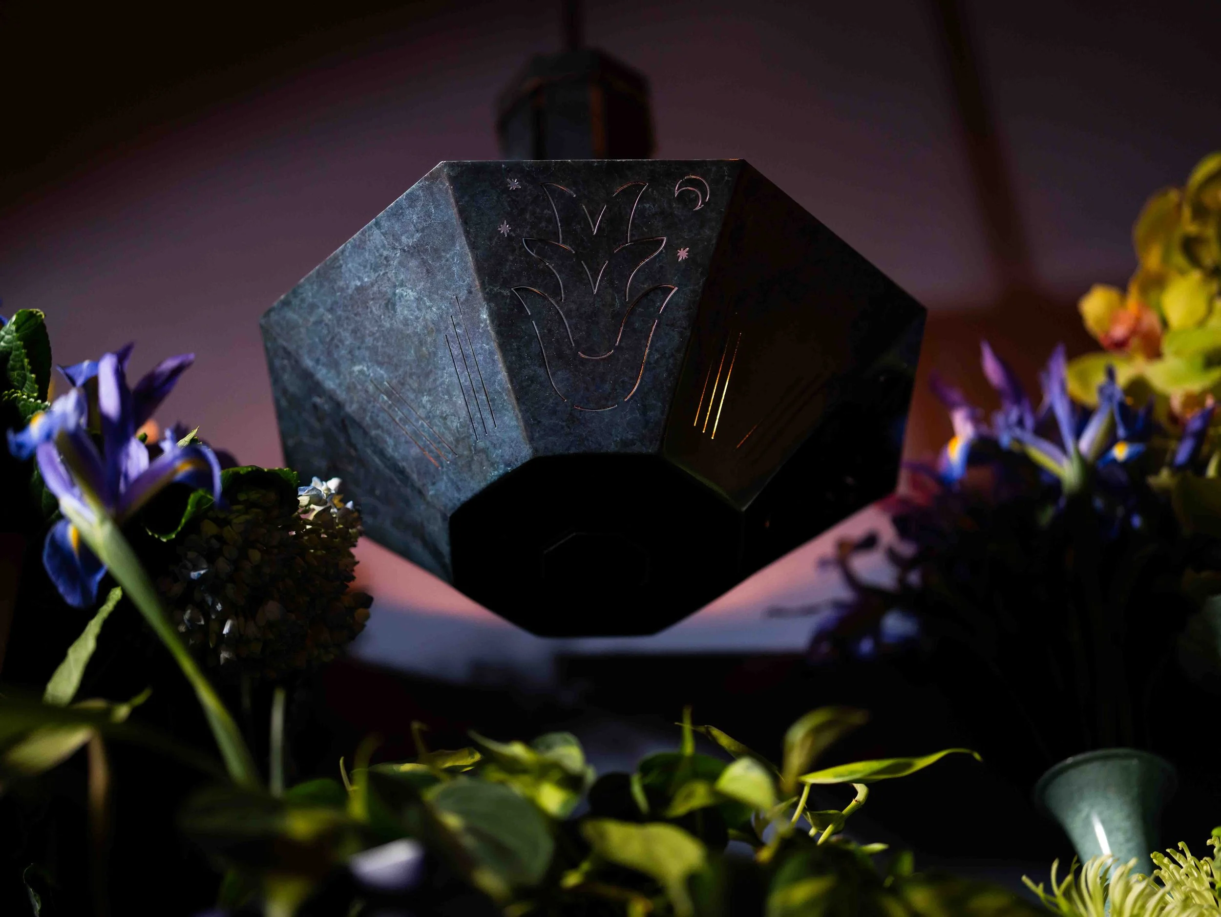 A decorative lamp hanging from the ceiling, illuminated with a warm light, surrounded by colorful flowers and greenery.