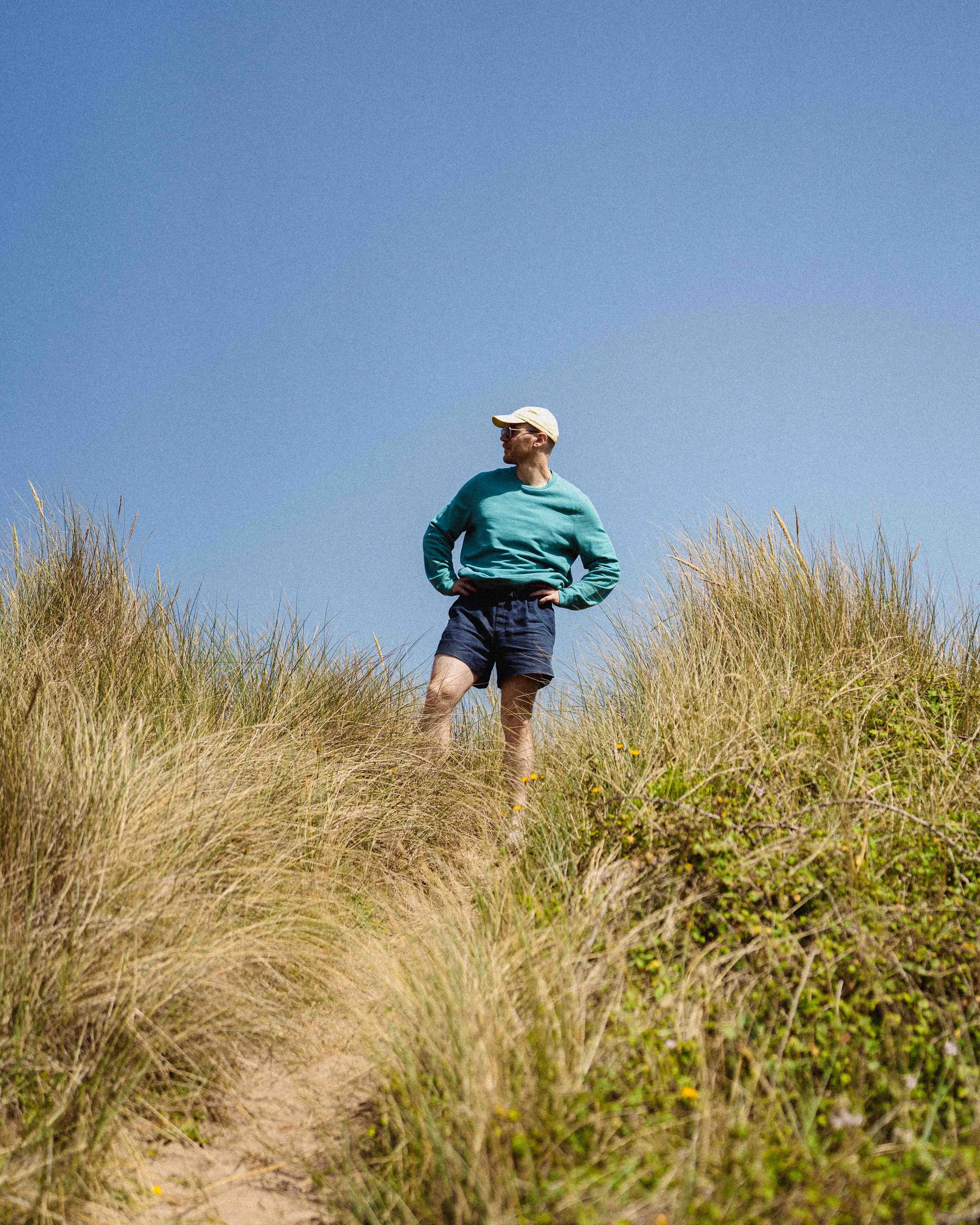 Person wearing sunglasses, a light-colored cap, turquoise jacket, and navy shorts standing among tall grass under a clear blue sky.