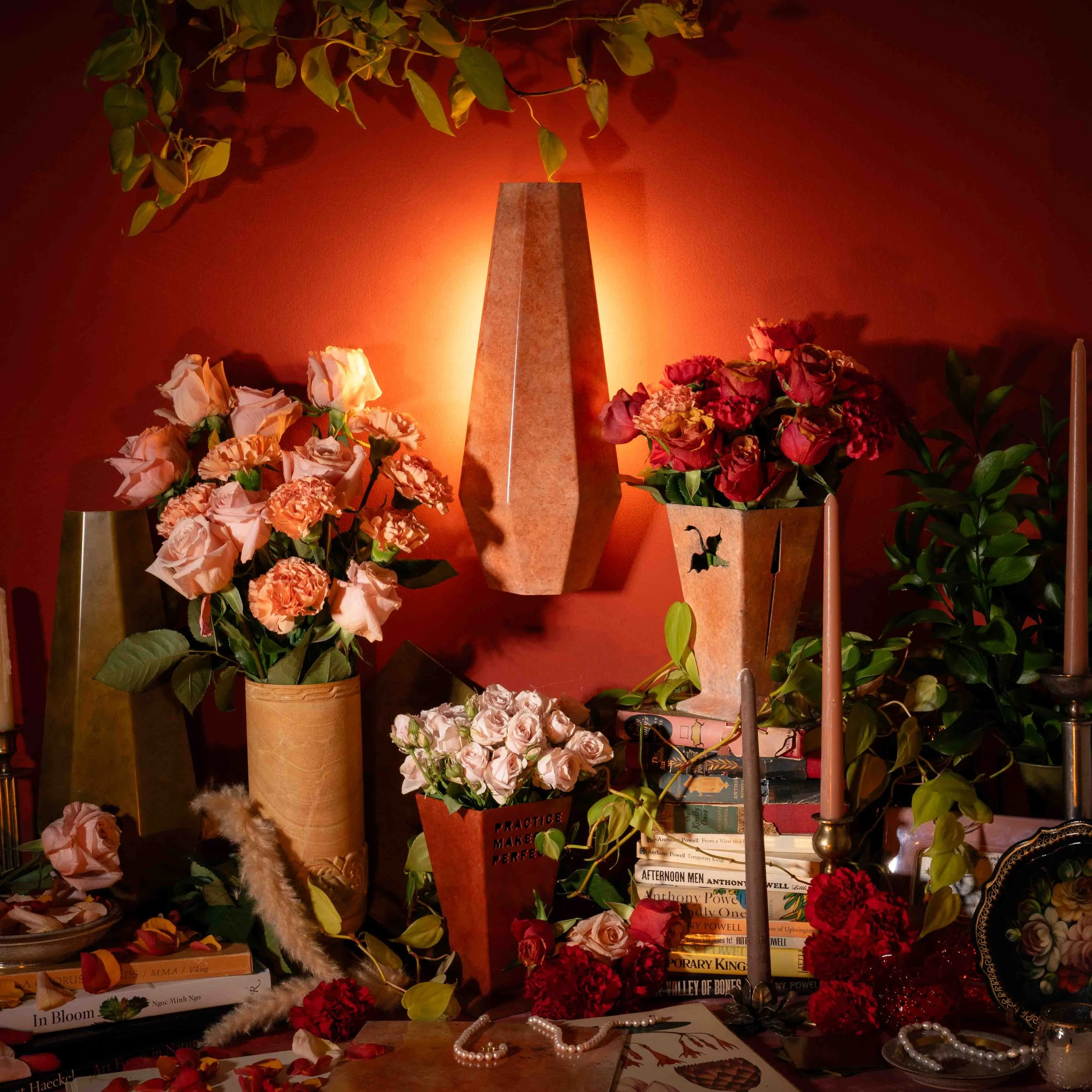 A shelf filled with various flowers and books, with a warm orange light illuminating the scene. There are vases of roses and other blossoms, a string of pearls, and a pink candle, with a red wall in the background.