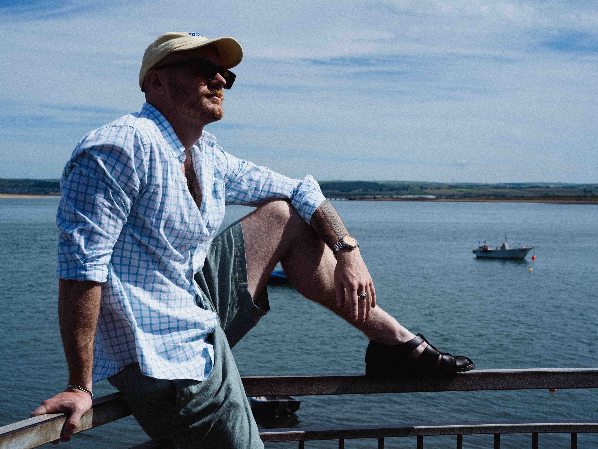 A man with a beard, wearing sunglasses, a beige cap, and a light checkered shirt, sitting on a railing by the water, with a boat and distant shoreline in the background, during daytime.