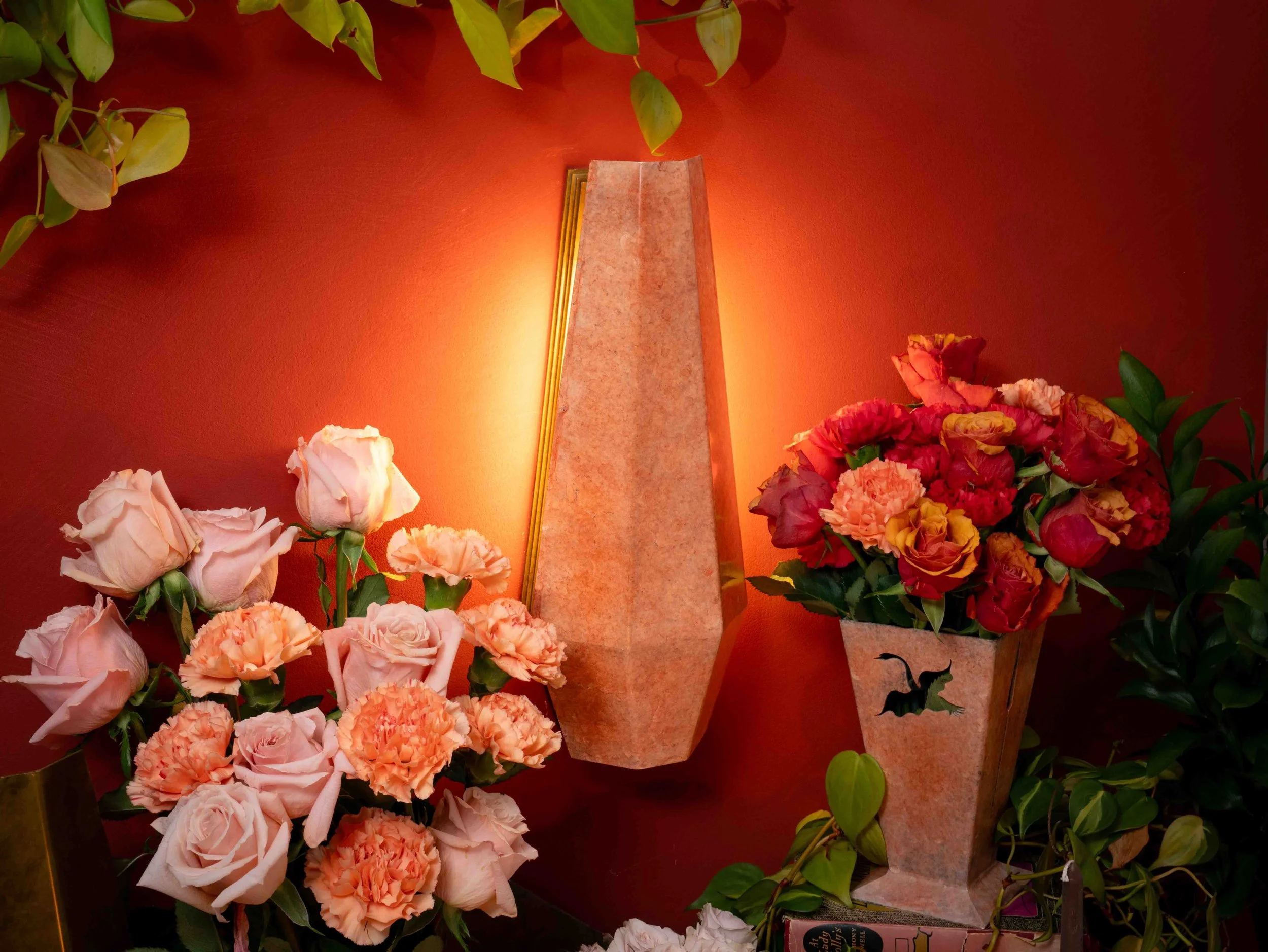 Pink and red carnations with green leaves surrounding a marble memorial with gold trim, illuminated against a red wall, likely in a memorial or funeral setting.