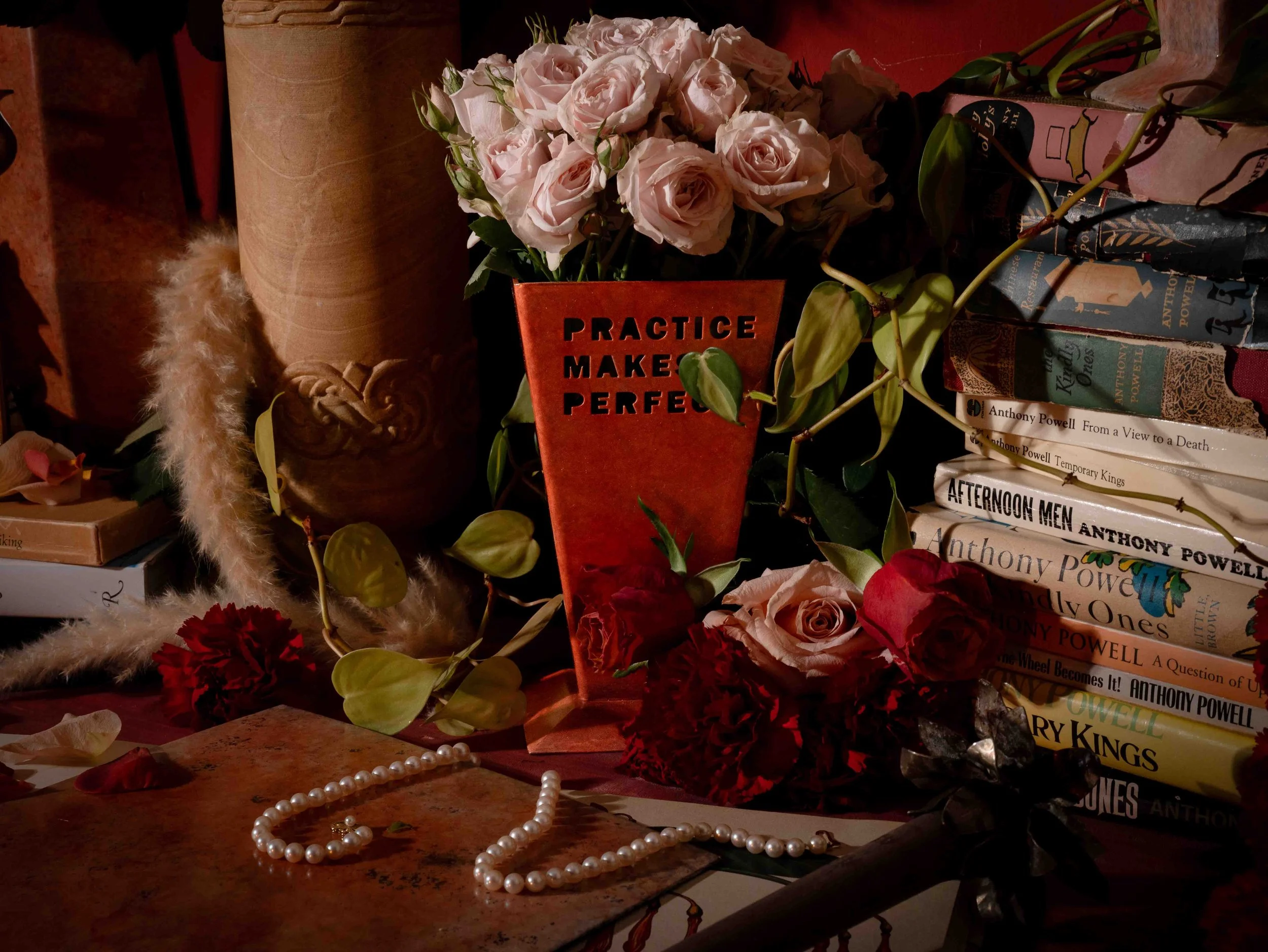 A cozy scene with a bouquet of light pink roses in a red container, a string of pearls, and a background of books and decorative items. The sign on the container reads "PRACTICE MAKES PERFECT."