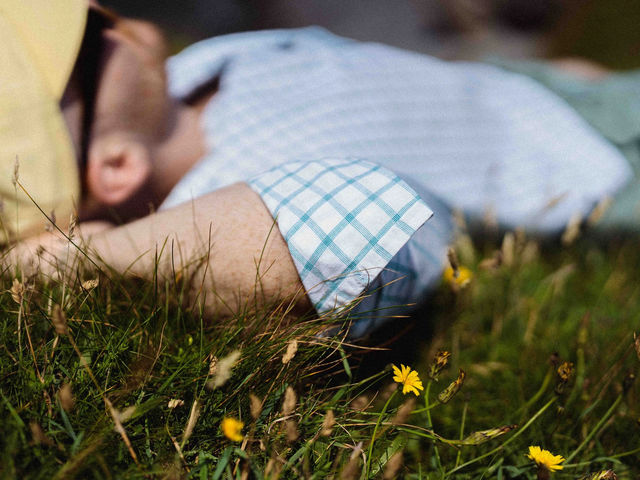 A child lying on the grass in a field with small yellow flowers, wearing a light blue checked shirt, with part of their face near the ground and eyes closed.