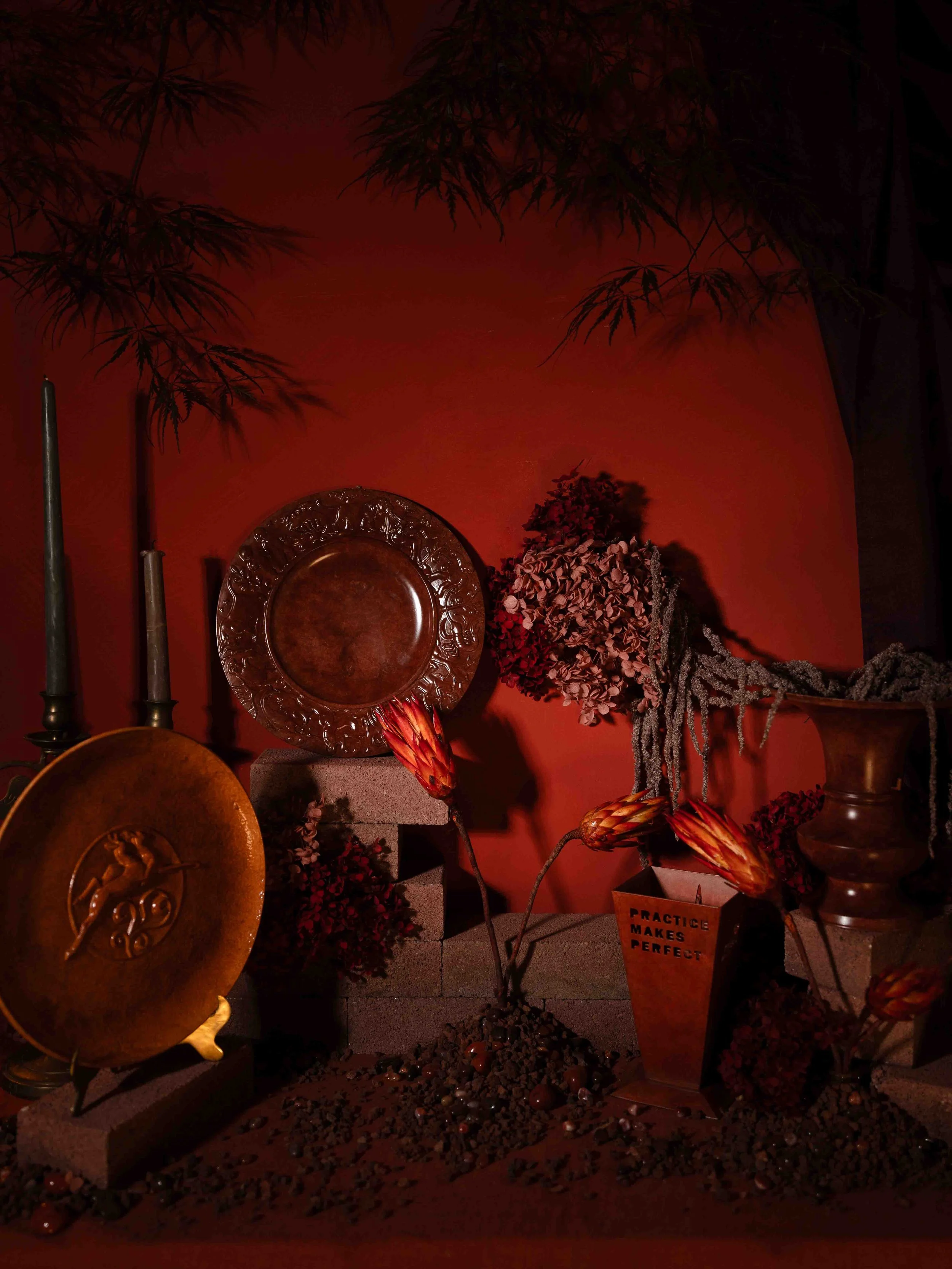 Decorative display with flower arrangements, large decorative plates, and vases against a red wall. Includes a small box with the message 'Practice makes perfect'.