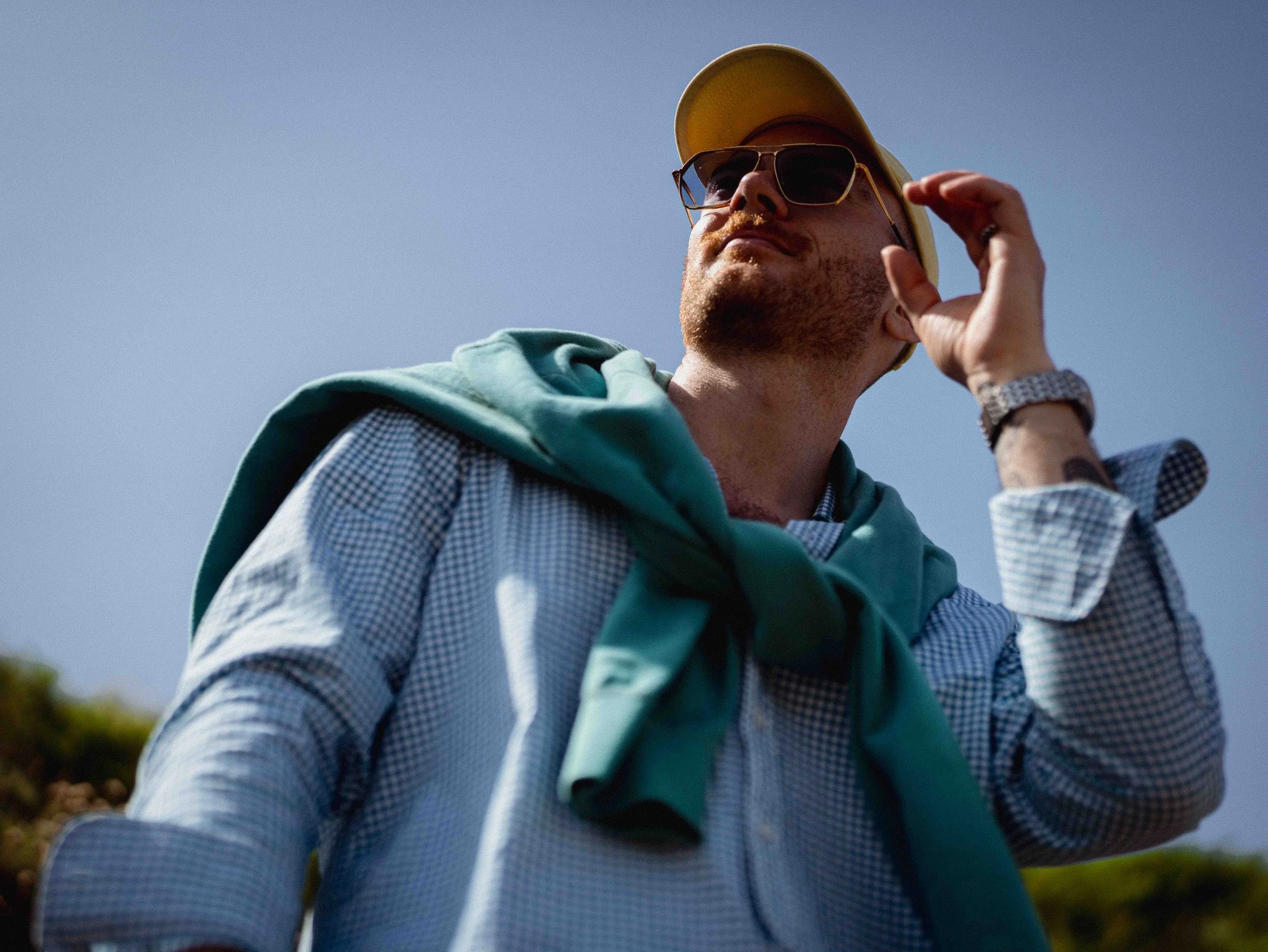 A man with sunglasses and a yellow cap adjusting his glasses outdoors against a clear sky.