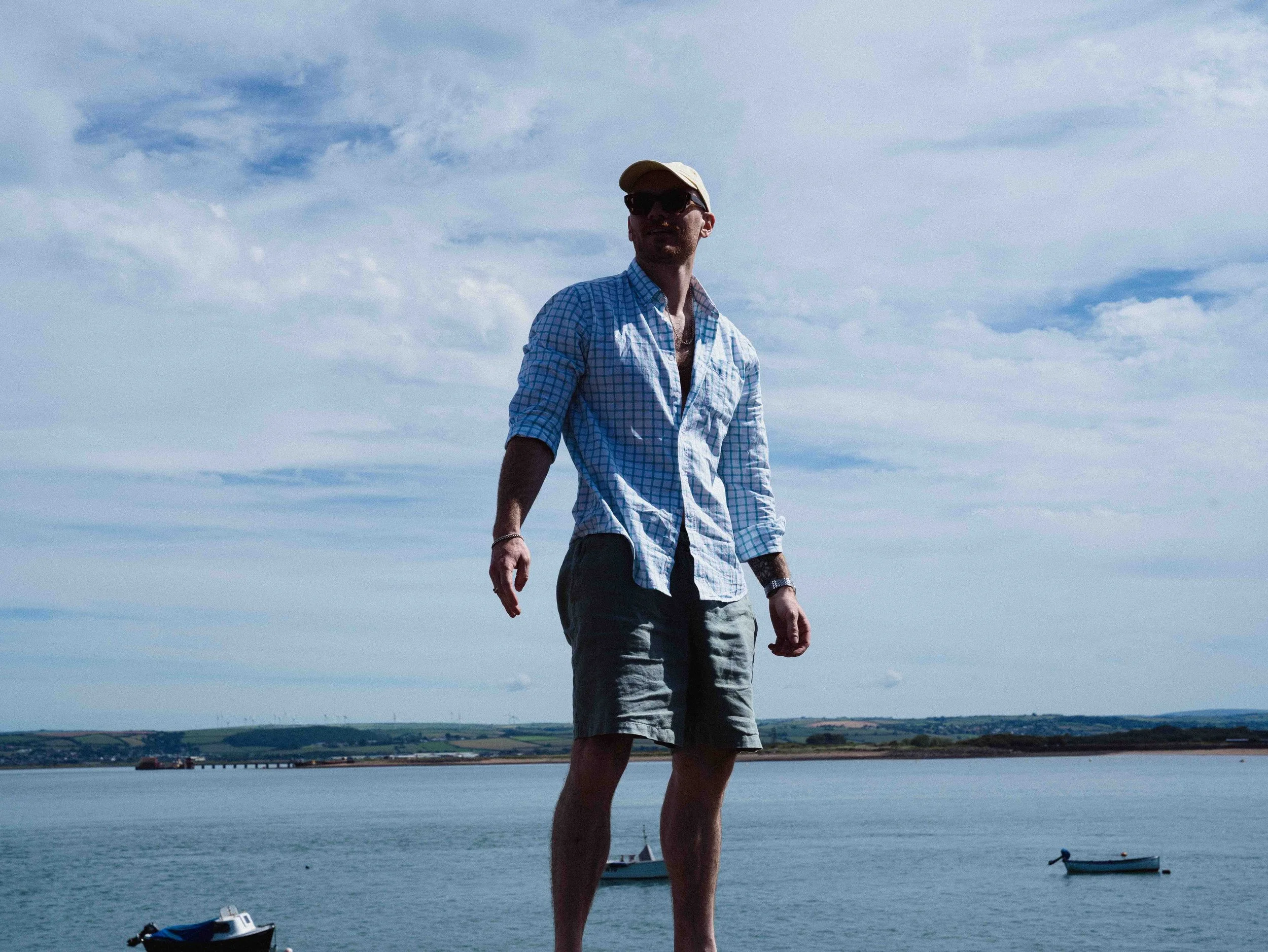 A man wearing a striped blue and white shirt, khaki shorts, sunglasses, and a beige cap standing by a body of water with boats, a lake, and hills in the background under a partly cloudy sky.