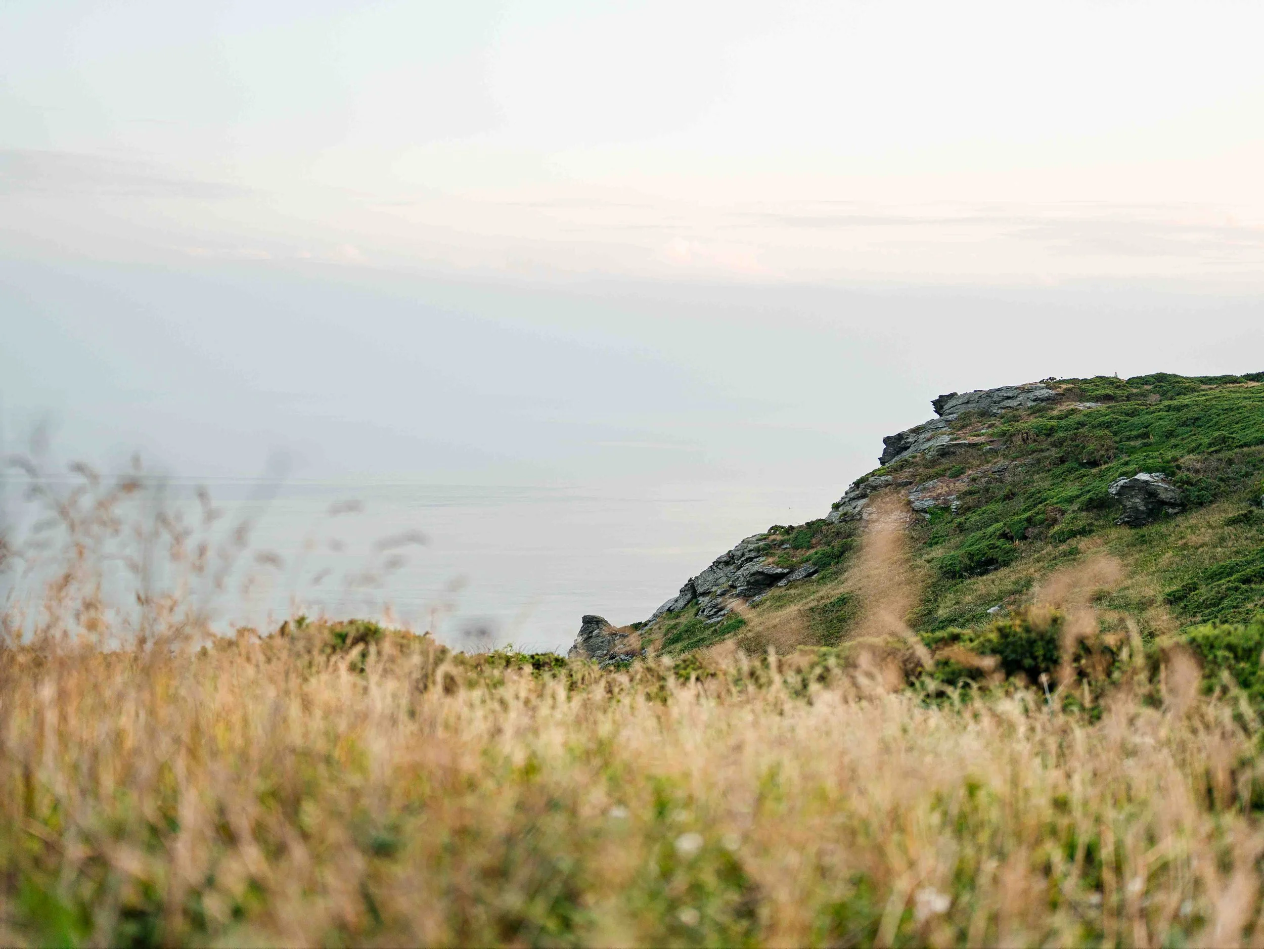 A grassy hillside with a rocky cliff on the right, overlooking a body of water with a cloudy sky.