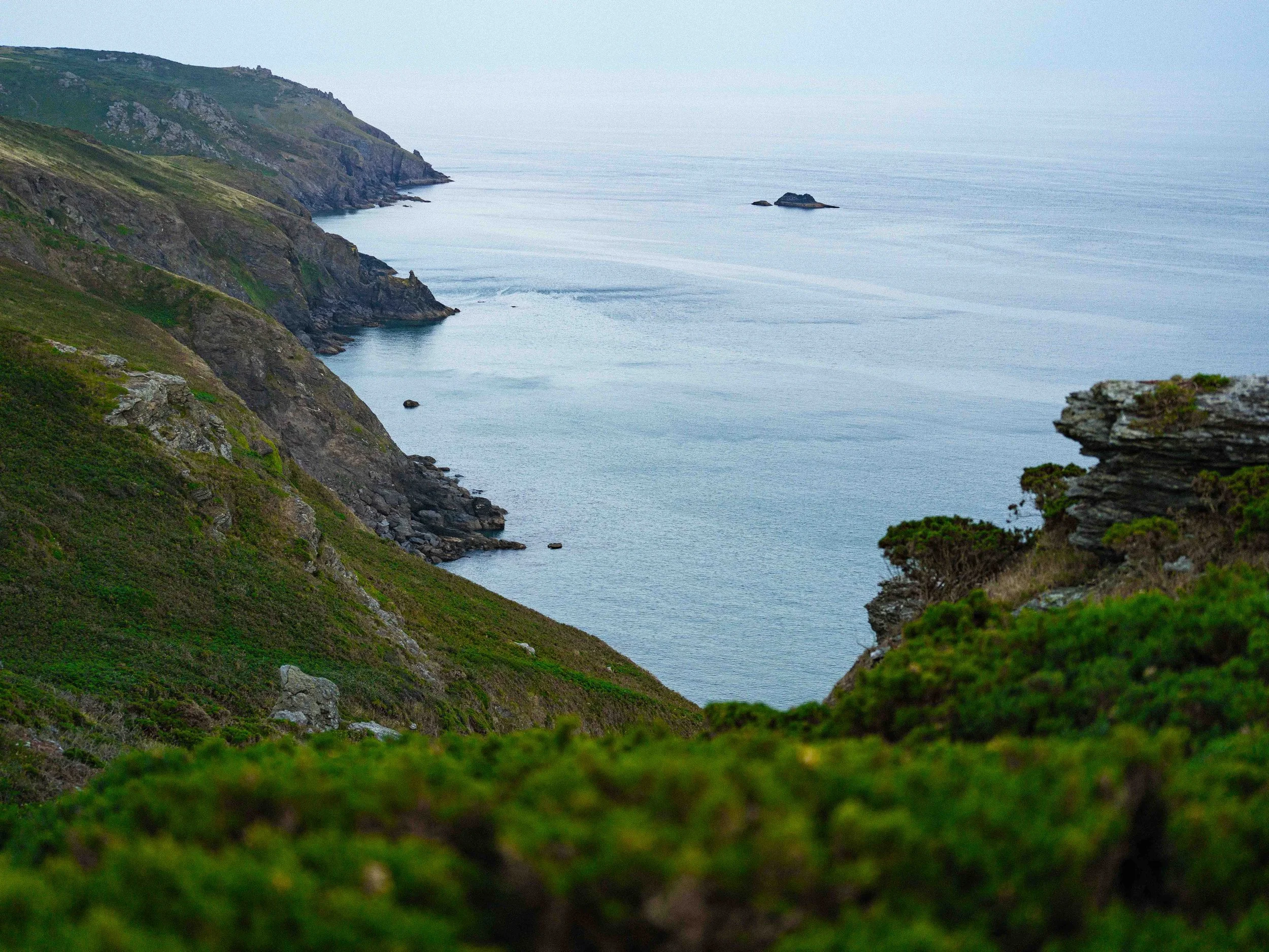 Coastal cliffs with green vegetation and rocks overlooking the calm ocean under a cloudy sky.