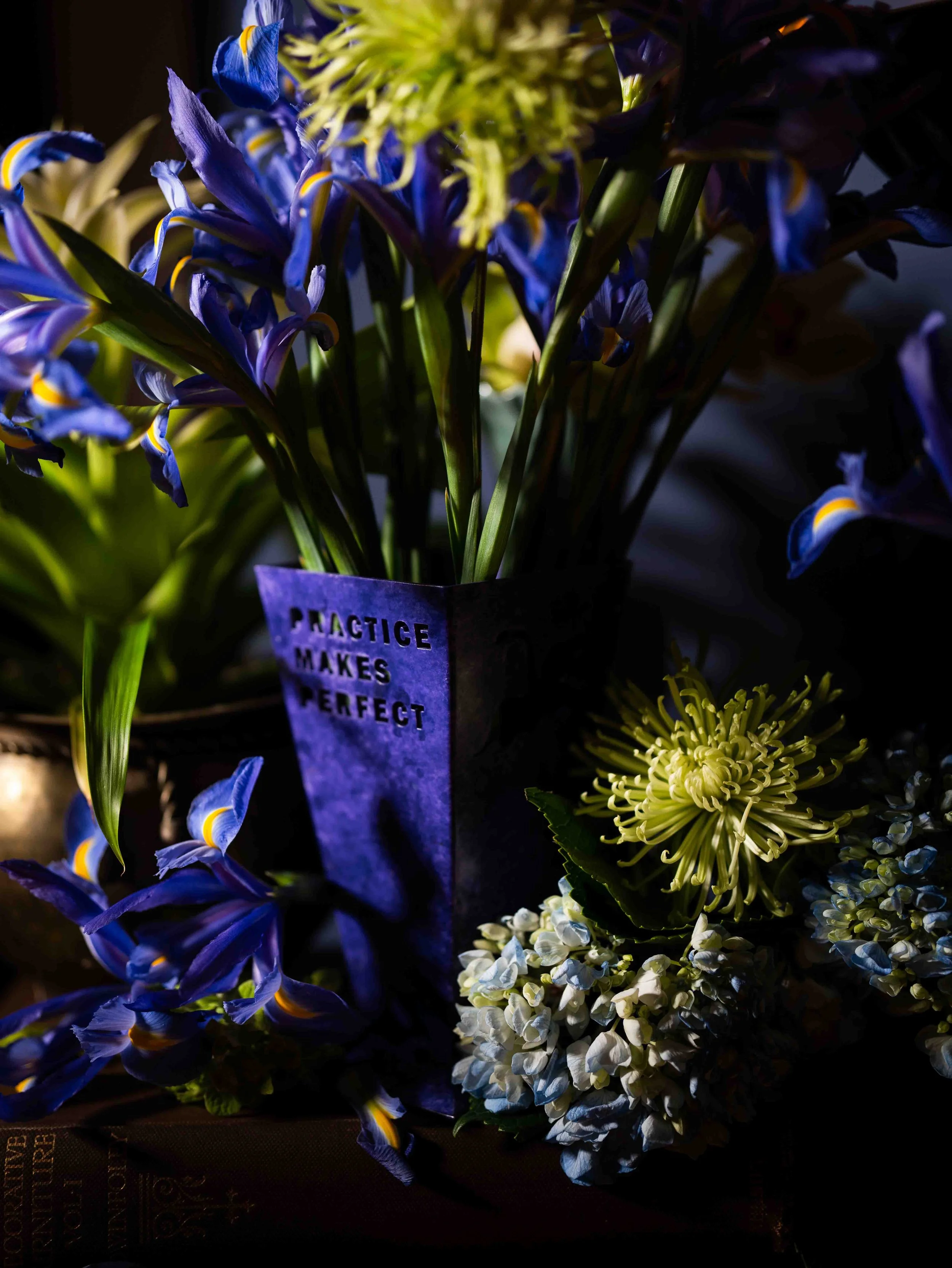 Close-up of a vibrant bouquet of blue and white flowers, with a card that reads 'Practice makes perfect' partially visible.