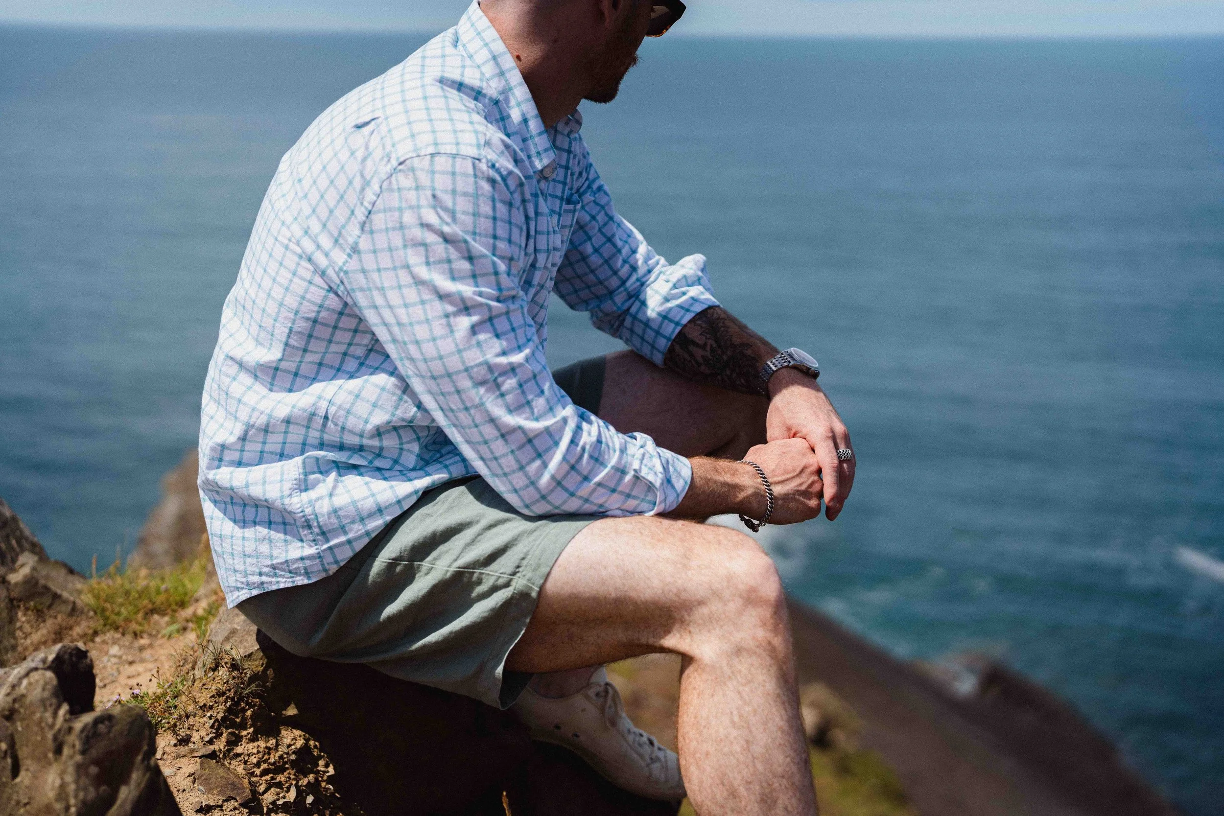 A man sitting on a rocky ledge by the ocean, wearing a checkered shirt, shorts, a watch, and a bracelet, looking down at his hands with a tattoo on his arm.