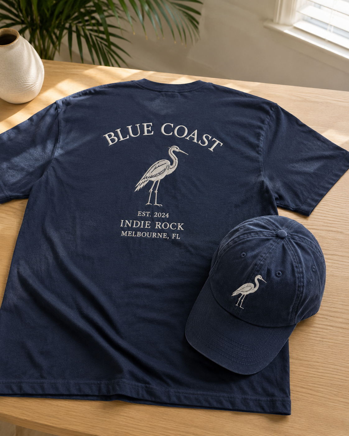 A navy blue T-shirt and matching cap with a heron logo, placed on a wooden table near a potted plant and a window.