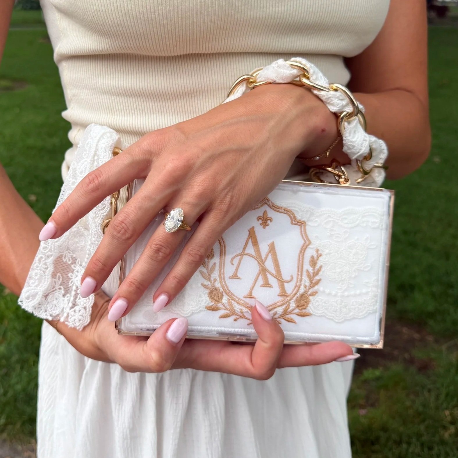 Close-up of a woman's hand holding a decorative box with gold embroidered initials  'A', inside a ribbon and lace bracelet, wearing an engagement ring with a large oval diamond. She is wearing a white sleeveless dress, standing outdoors on grass.