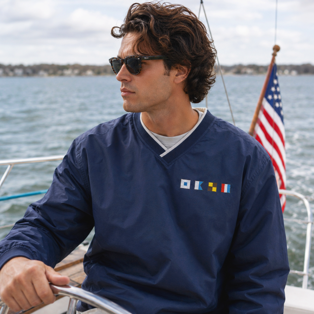 Man with wavy brown hair wearing sunglasses and a navy blue jacket with nautical patches, steering a boat on a body of water with American flag in the background.