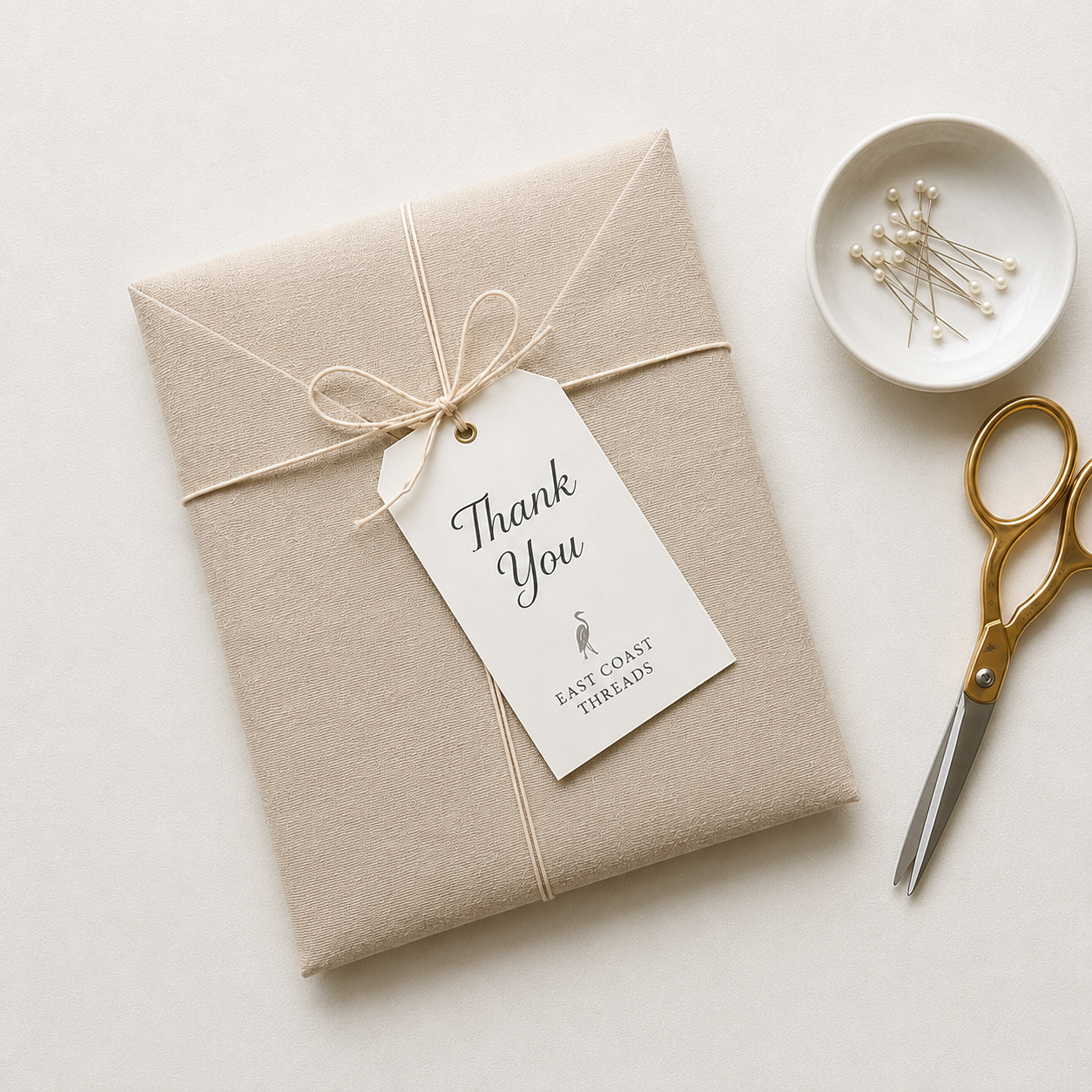 A wrapped gift with a 'Thank You' tag from East Coast Threads, placed on a white surface alongside gold-handled scissors, a small white dish with pearl-headed pins, and white string.