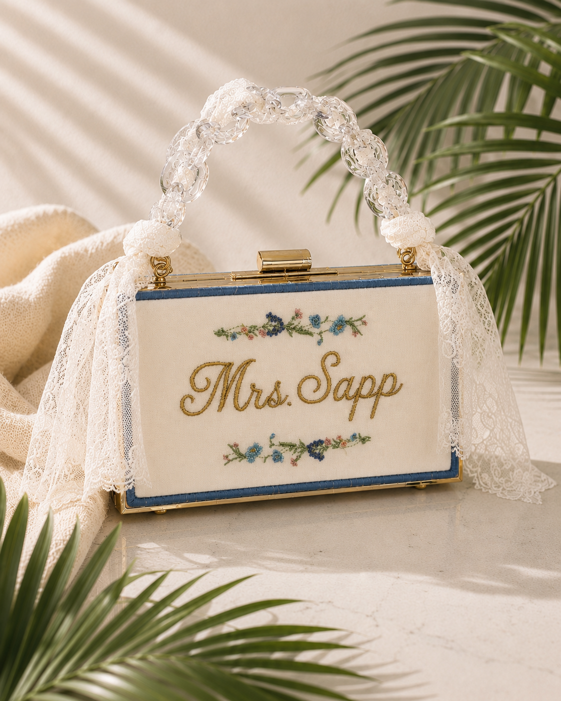 A vintage clutch purse with embroidered floral design and the name "Mrs. Sapp" on the front, decorated with lace, pearls, and rhinestones on the handle, surrounded by tropical leaves.