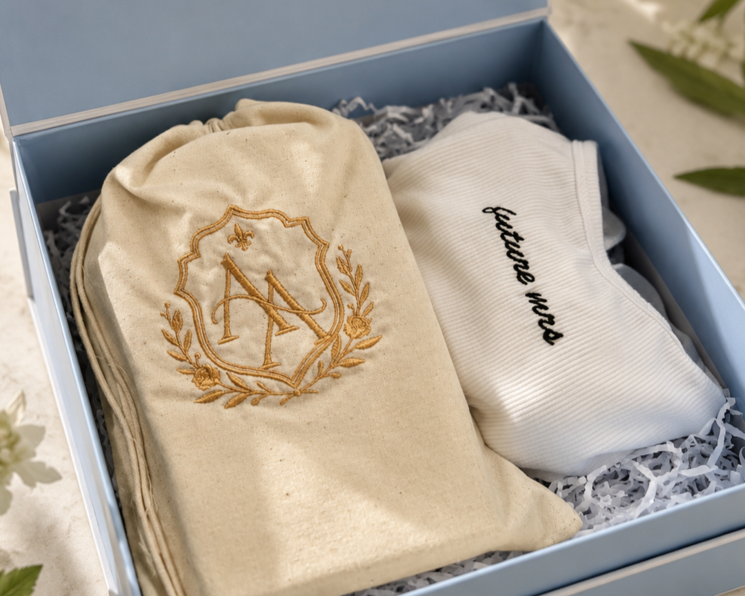 A person working on a fabric tote bag design with a sketchbook, pen, fabric, and sewing supplies on a table. There are folded clothes with 'East Coast' embroidered on the top, a plant, and a measuring tape.