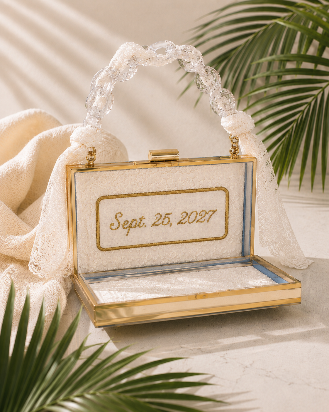 An elegant glass jewelry box with a gold frame, lace interior, and a handle made of clear plastic links. The inside of the box has the date "Sept. 25, 2027" embroidered in gold on a lace background, set against a neutral fabric with decorative lace and green palm leaves in the backdrop.
