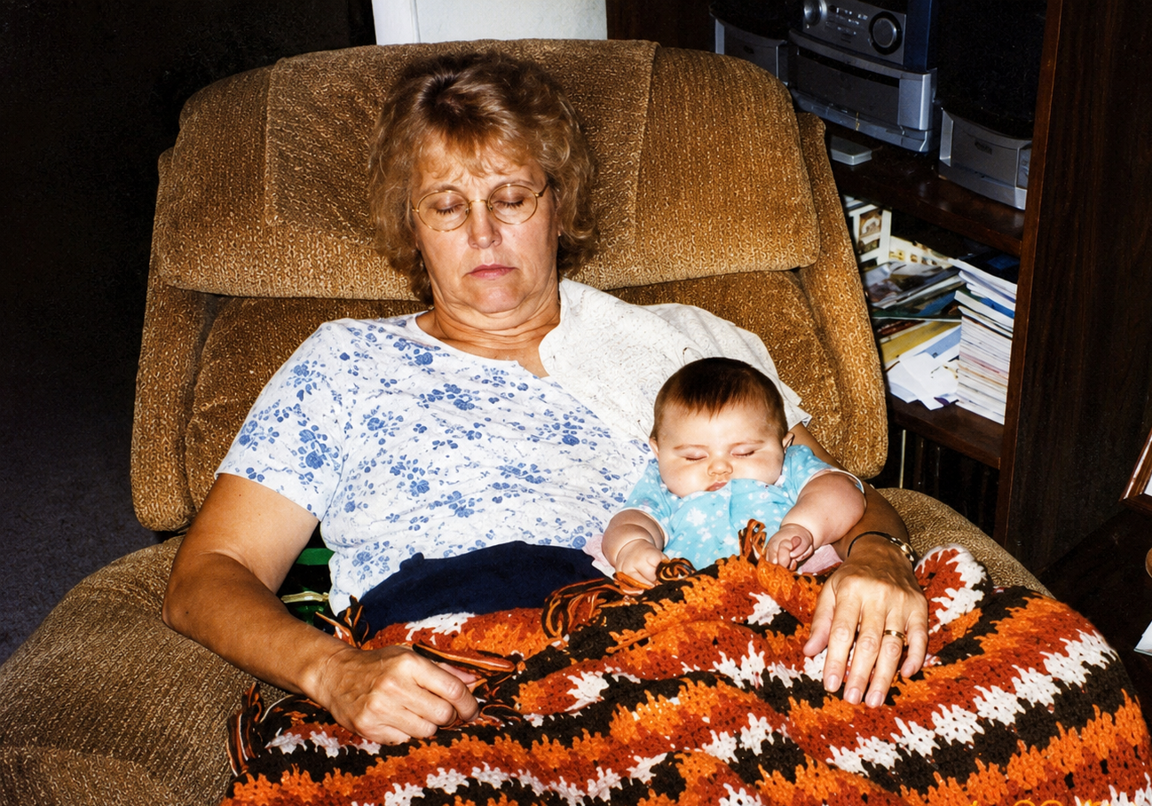 An elderly woman with glasses sleeping in a brown armchair with a baby resting on her lap, covered with a multicolored crocheted blanket.