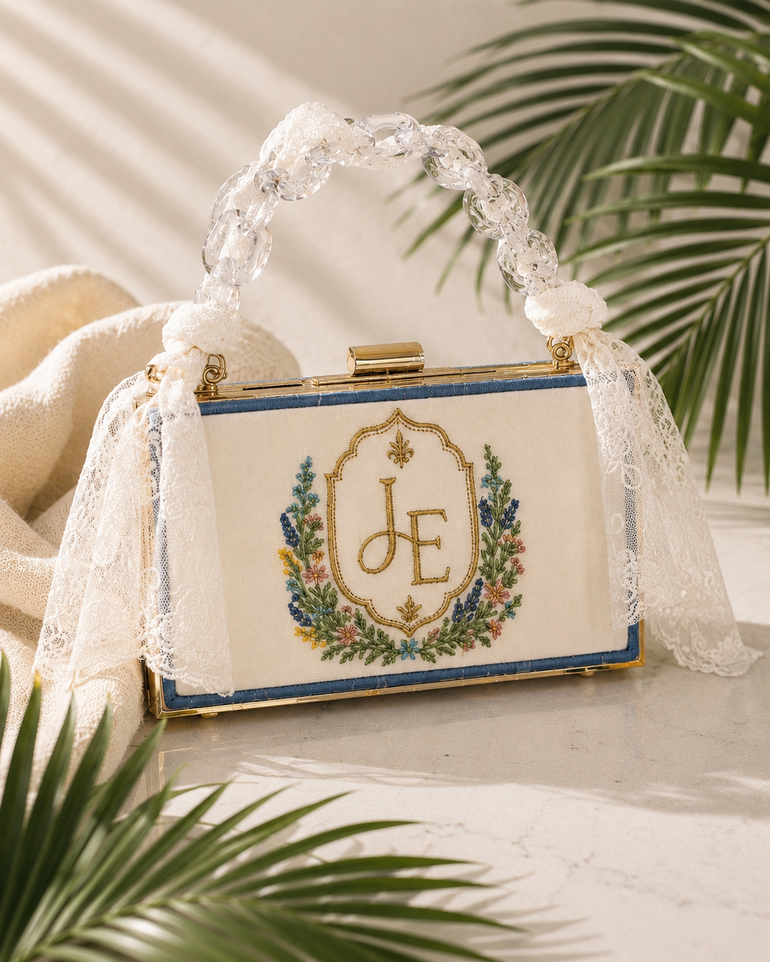 A vintage embroidered handbag with monogram initials J E, decorated with a floral wreath, lace, and a bead handle, placed on a marble surface next to a beige fabric and a green palm leaf.