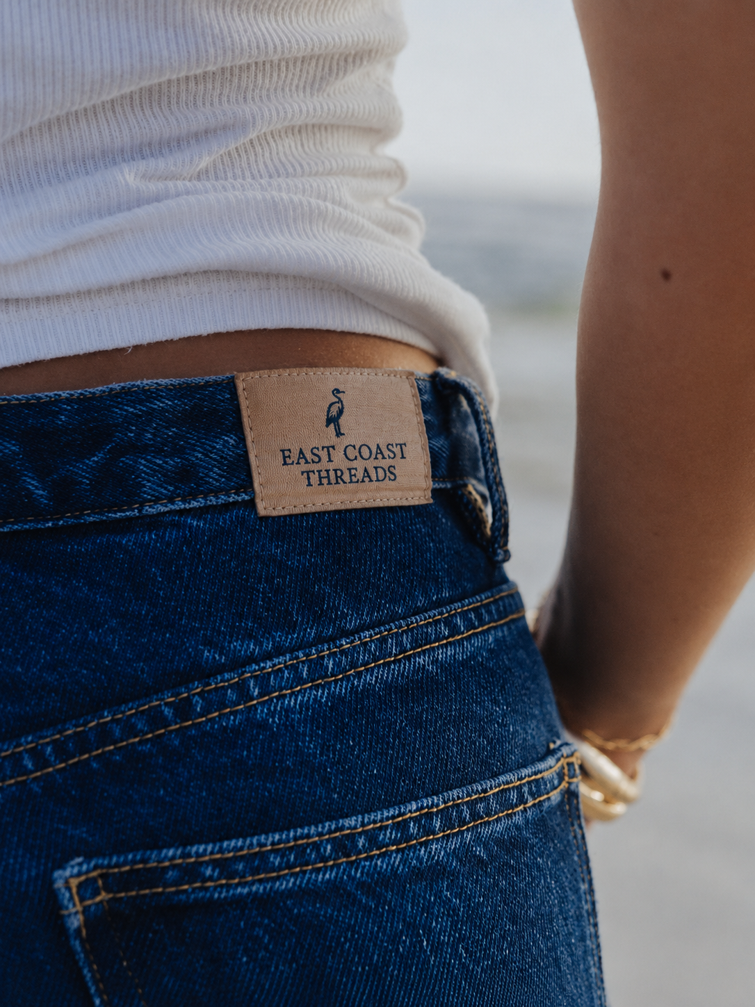 Close-up of a person wearing blue jeans with a beige label that says "East Coast Threads" and a white top, standing near the beach.