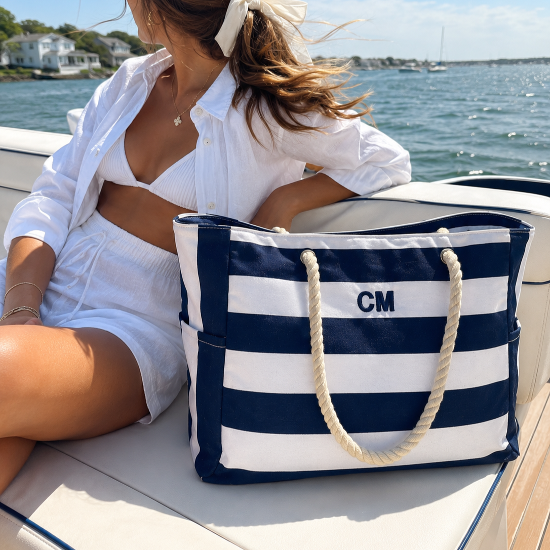 A woman sitting on a boat near a body of water, wearing a white bikini top, shorts, and a white shirt, with a blue and white striped tote bag that has the initials 'CM' on it.