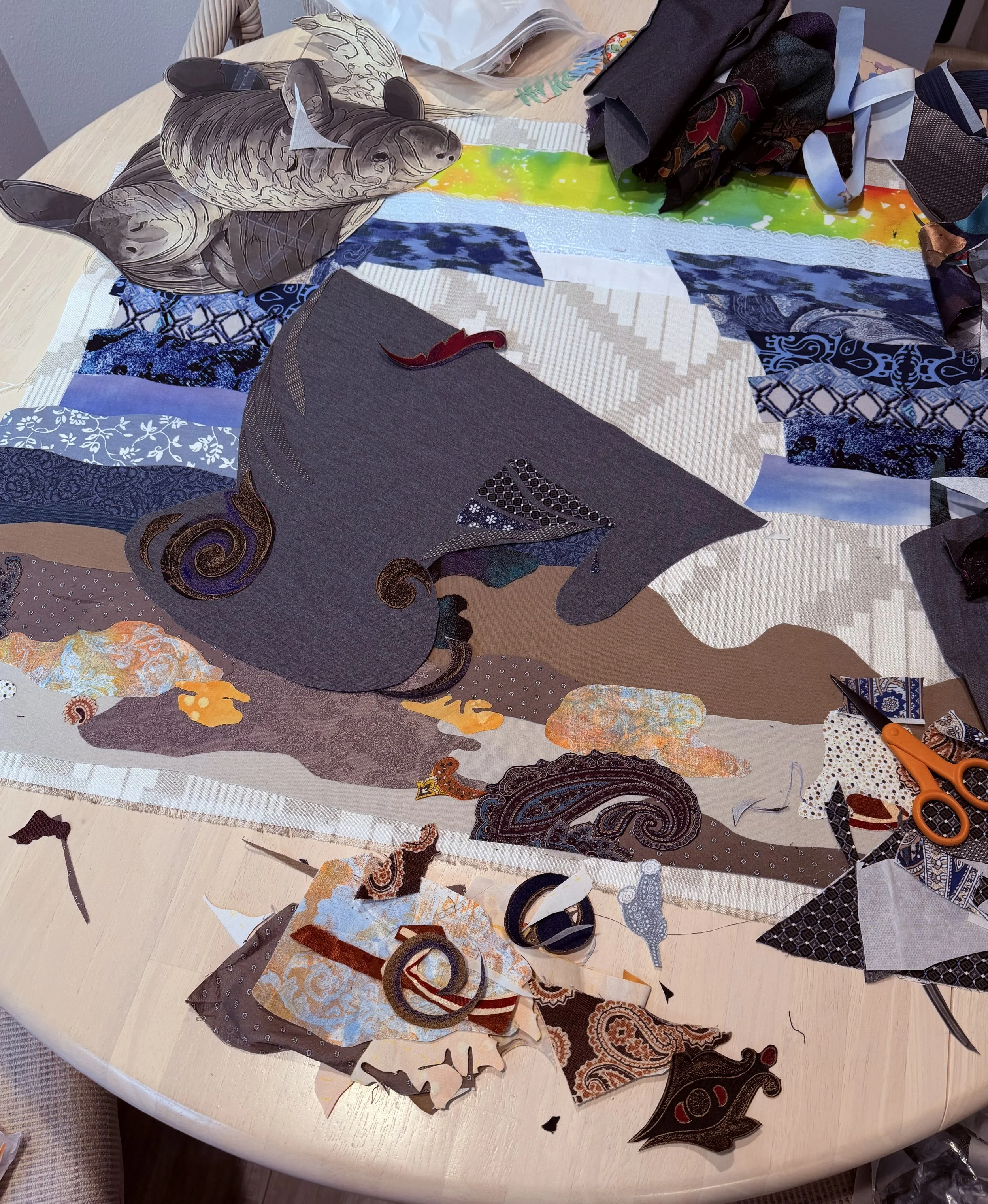 Various fabric cutouts and scraps scattered on a round wooden table, including a fabric with a horse face and intricate patterns, scissors, and torn pieces of cloth.