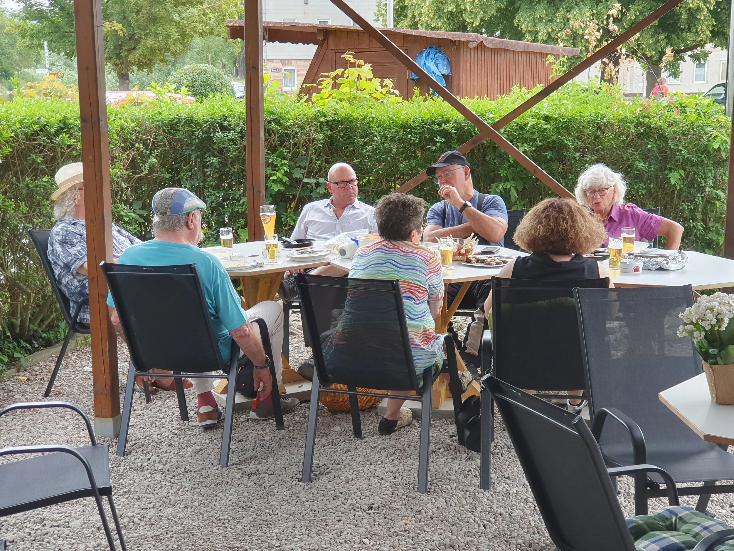 Ein Garten mit einem gedeckten Tisch, auf dem Essen und Getränke für mehrere Personen stehen. Es sitzen mehrere ältere Menschen und eine jüngere Person an dem Tisch, sprechen und genießen das Essen im Freien. Im Hintergrund sind Bäume, eine Hecke und ein kleines Holzgebäude sichtbar.