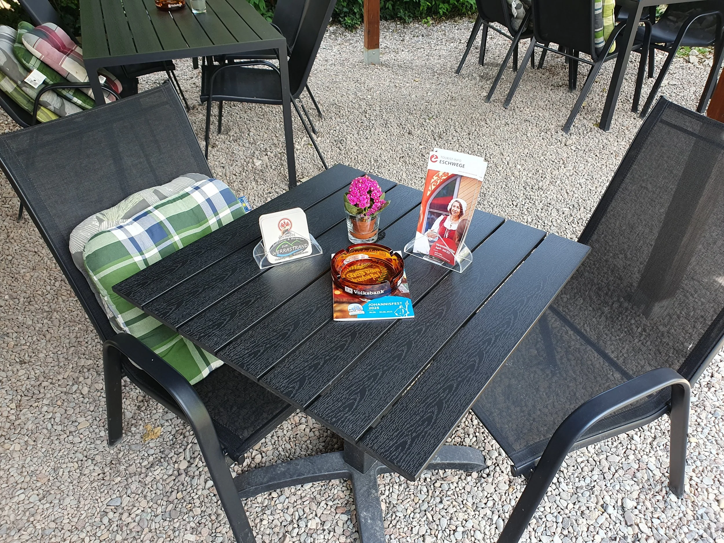 Ein graues Gartenmöbel-Set mit einem Tisch, mehreren Stühlen und einem Sitzkissen auf einem Kiesboden, im Hintergrund Pflanzen und weitere Stühle.