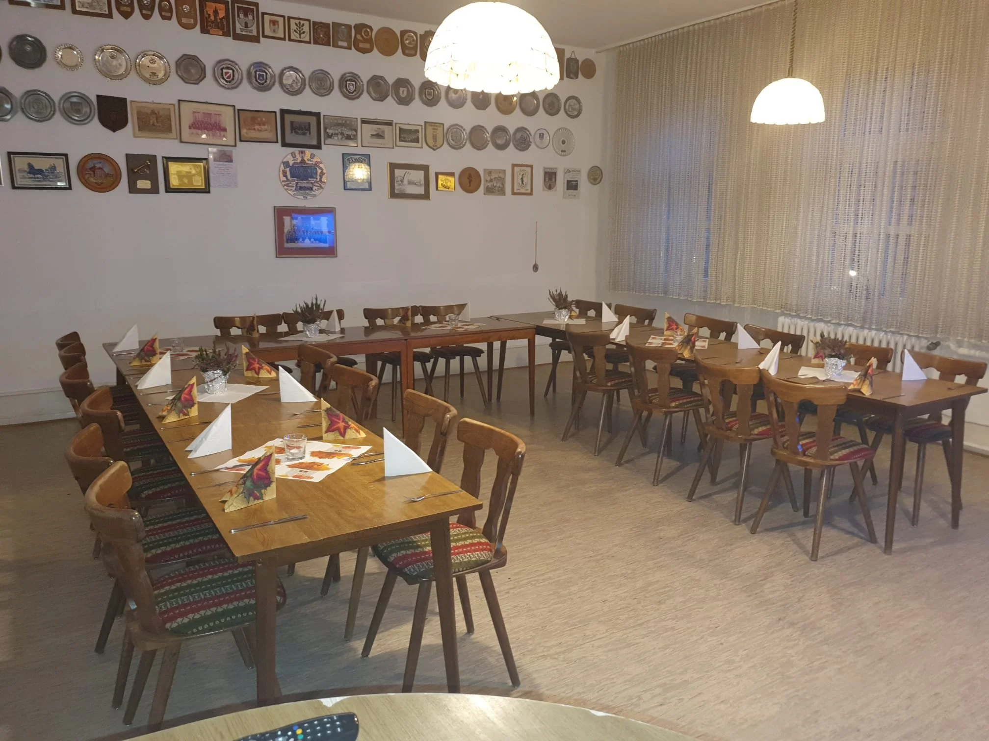 Ein Esszimmer mit mehreren Holztischen und Stühlen. An der Wand hängen verschiedene Trophäen, Bilder und Schilder. Drei Deckenleuchten hängen von der Decke. Es gibt lange Vorhänge vor einem Fenster.
