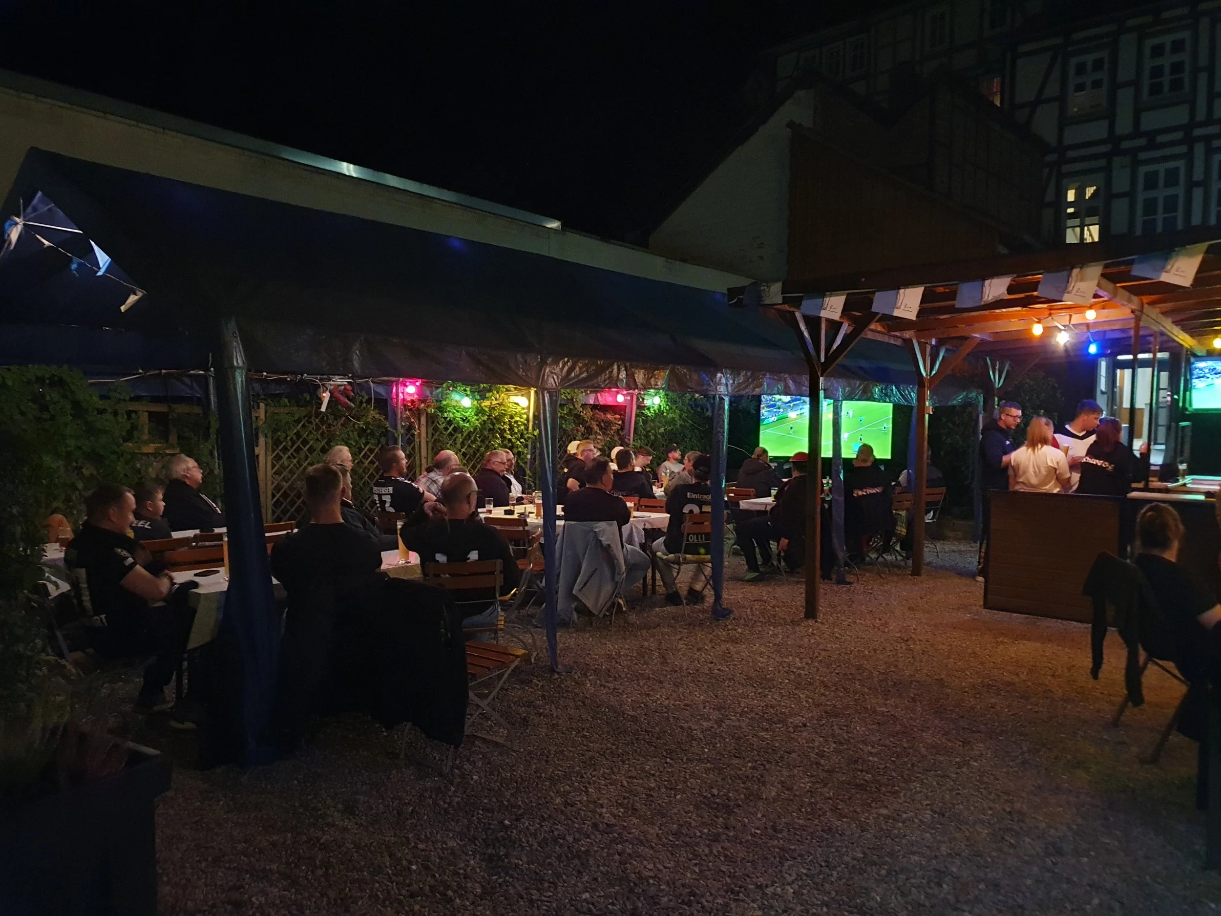Menschen sitzen im Freien und schauen Fußball auf einem großen Fernseher bei Nacht, gepflasterter Boden und bewaldeter Hintergrund.