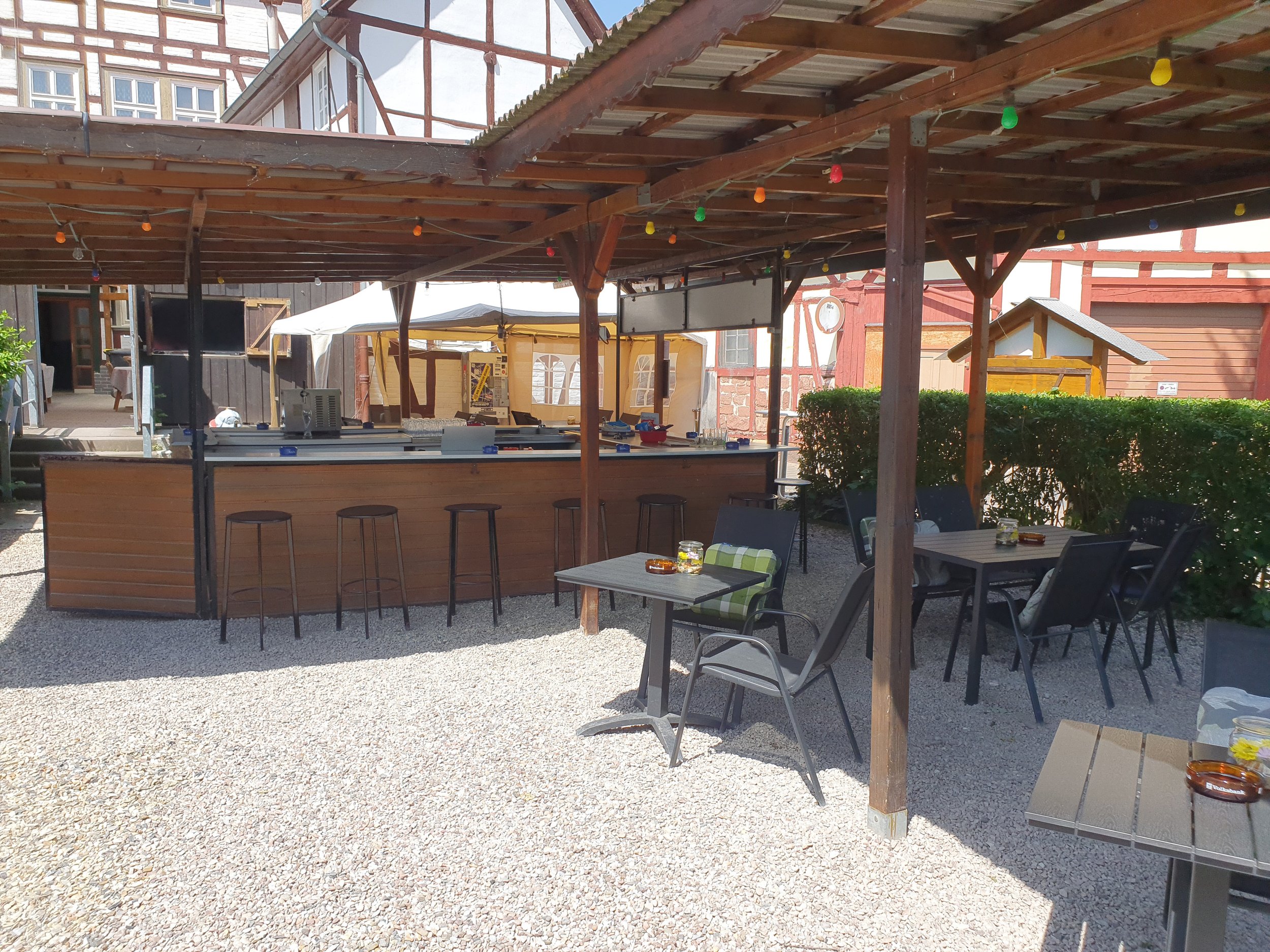Ein Außenbereich mit einer Bar im Vordergrund, mehreren Tischen und Stühlen, und einem Holzunterstand mit bunten Lichterketten hanging. Im Hintergrund sind alte Gebäude im Fachwerkstil sichtbar.