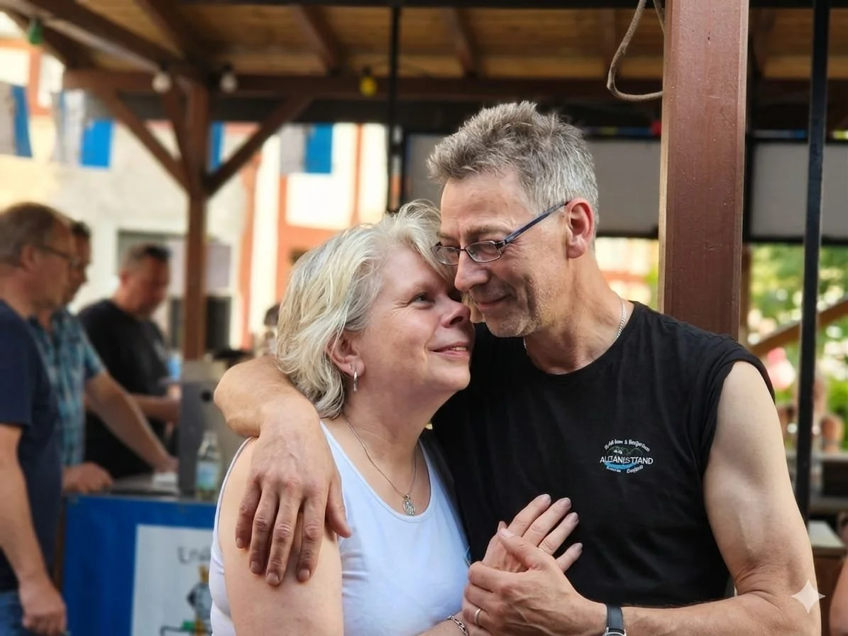 Ein Mann und eine Frau umarmen sich liebevoll und sehen sich in die Augen. Sie sind auf einer Veranstaltung im Freien, im Hintergrund sind weitere Personen und eine Bar-Counter sichtbar.