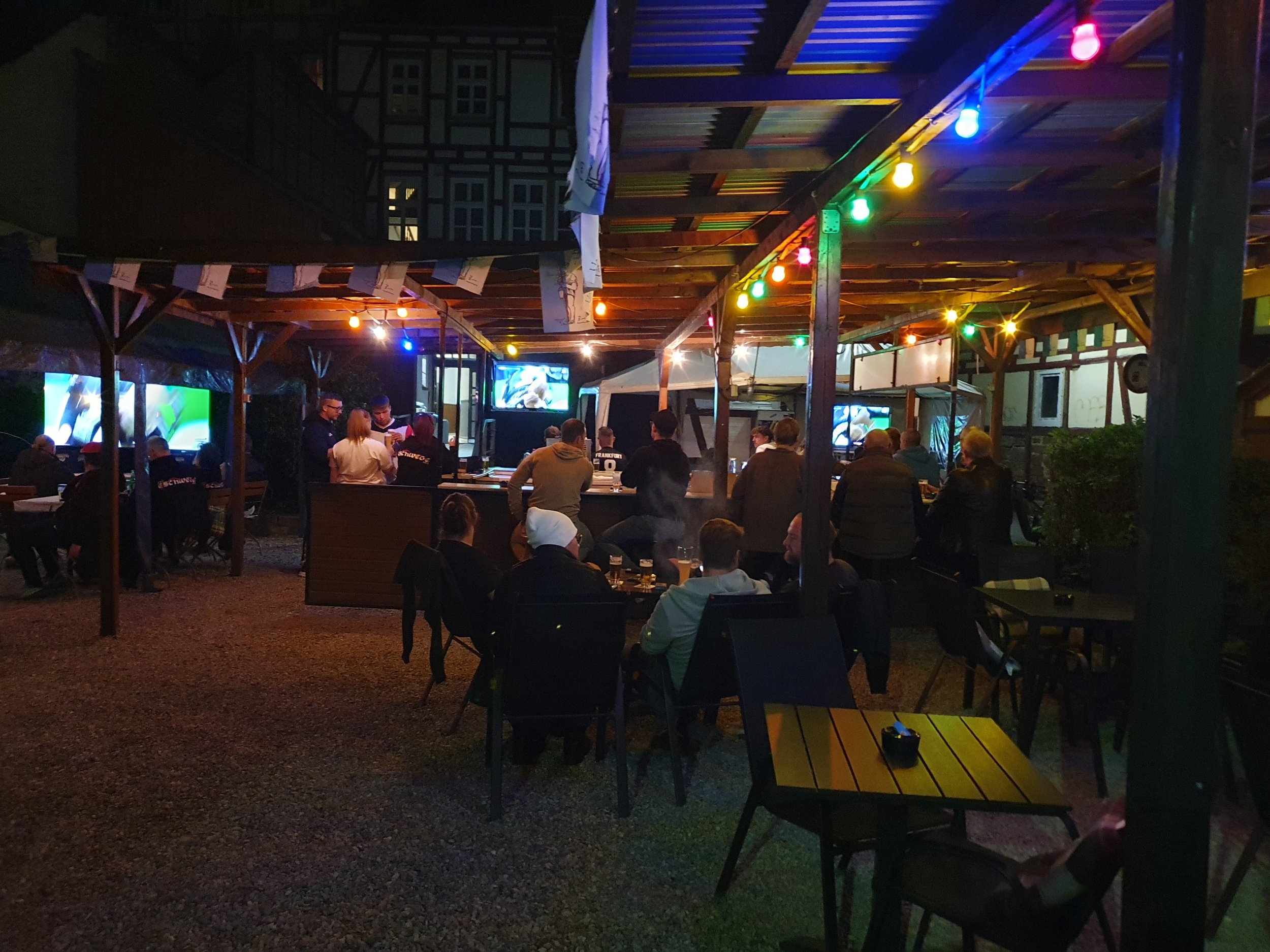Menschen sitzen abends in einer offenen Bar oder einem Gasthaus mit Holzbauten unter bunten Lichterketten, während Fernsehbildschirme im Hintergrund Fußball zeigen.
