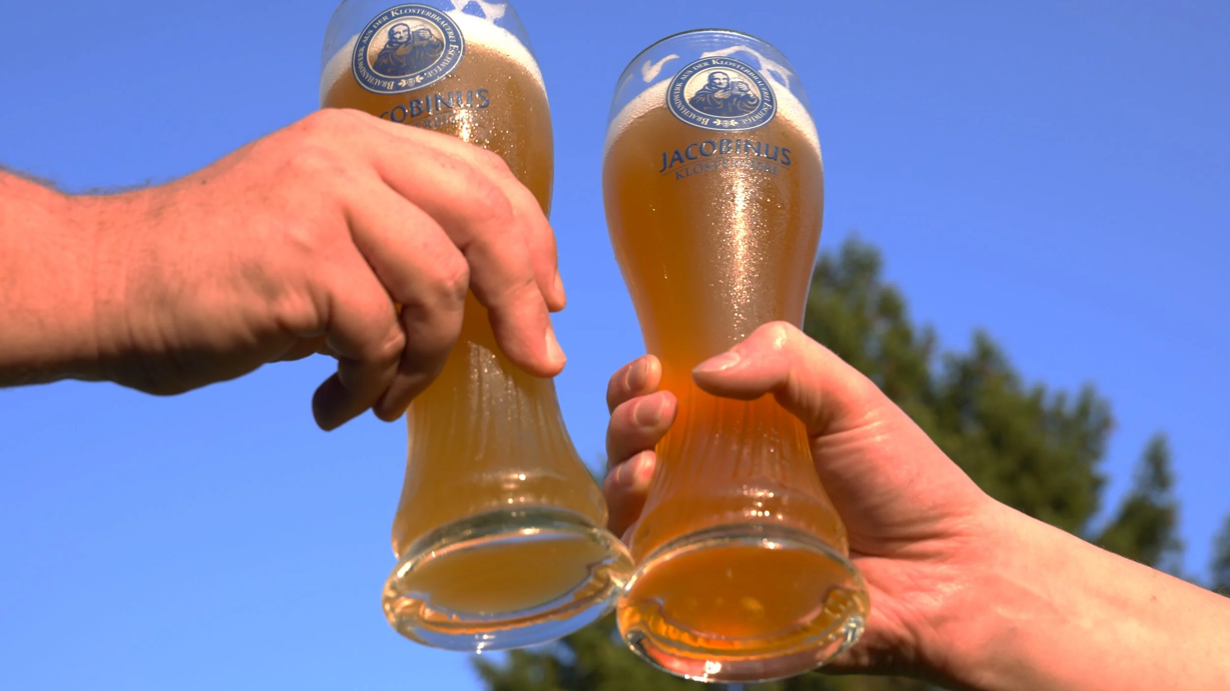 Mehrere Menschen stoßen mit Biergläsern an, die mit einer goldenen Biermischung gefüllt sind, gegen einen blauen Himmel im Hintergrund.