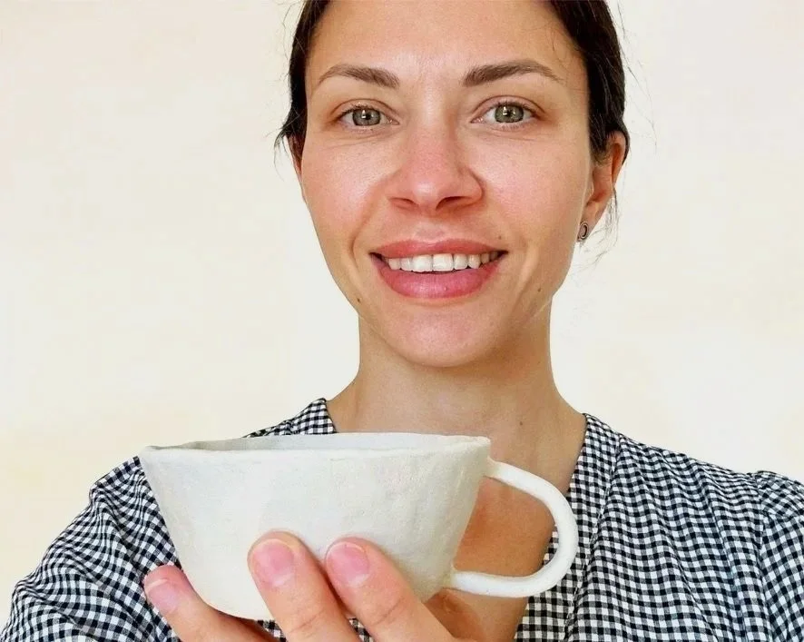 A woman with light skin and dark hair smiling while holding a large white handbuilt ceramic mug.