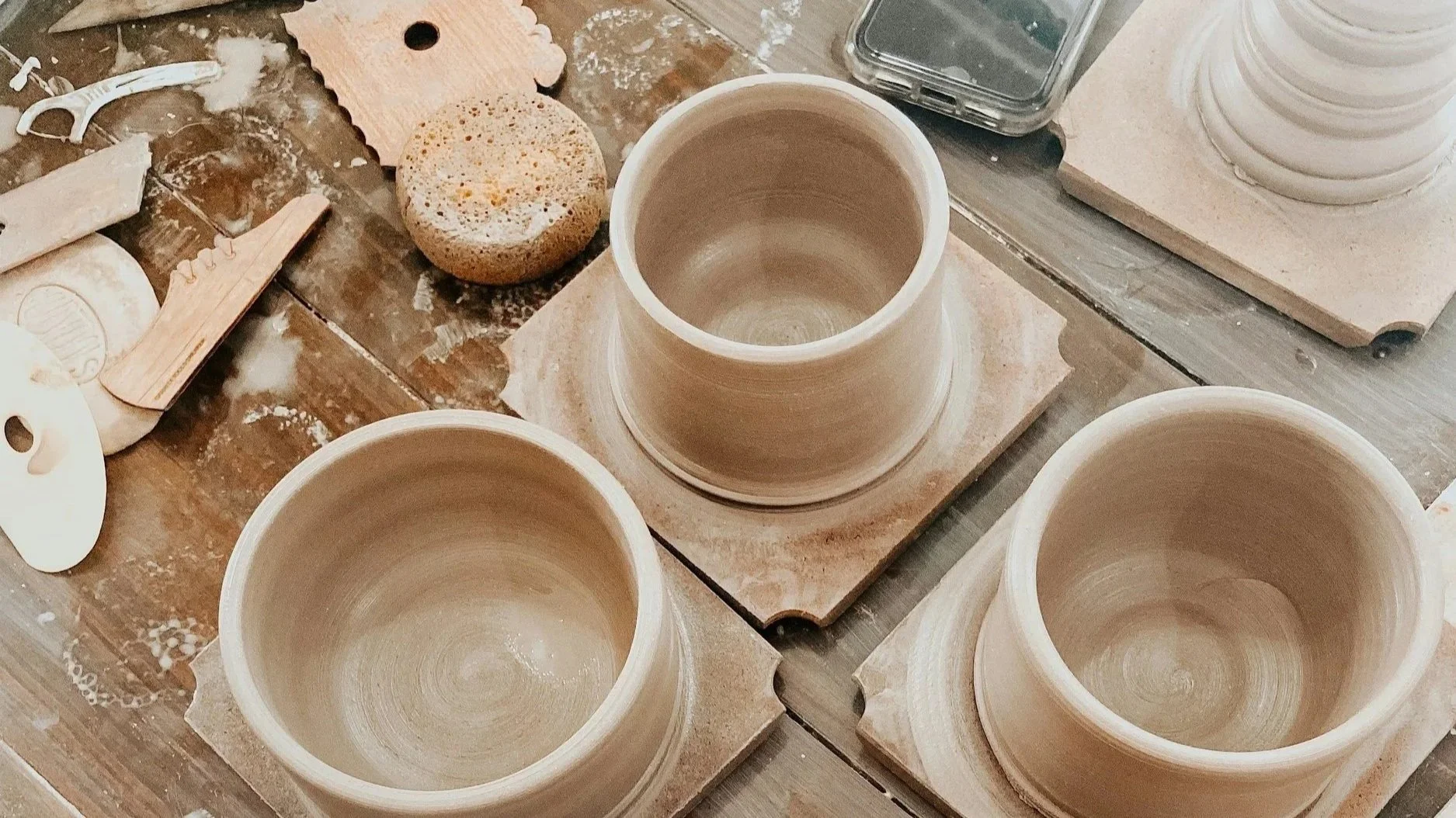 Three unfinished ceramic pots on a wooden worktable surrounded by pottery tools and supplies in a clay workshop.