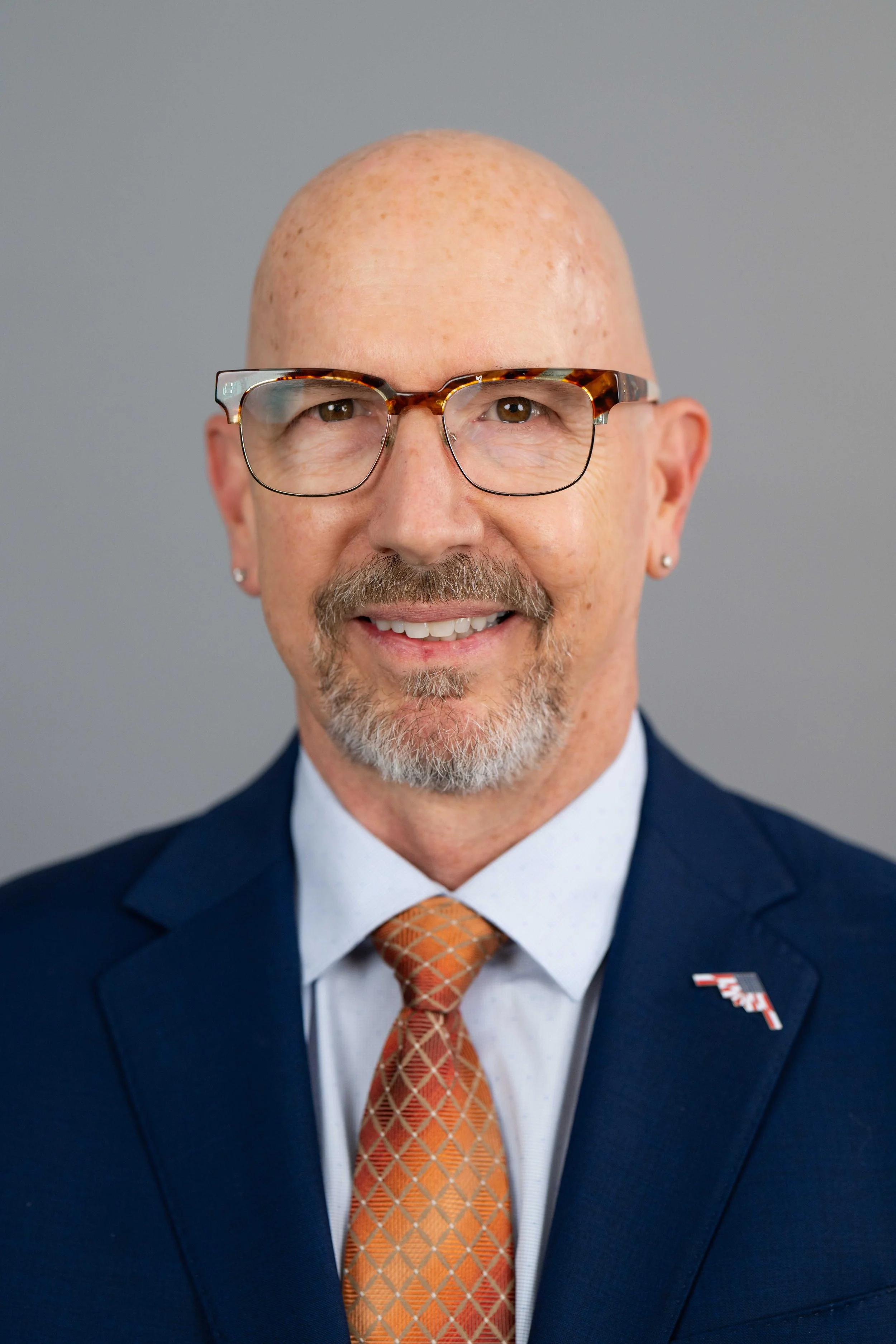 A man with a bald head, light brown eyes, and a goatee, wearing glasses, a navy suit, a white shirt, and an orange patterned tie, smiling against a gray background.