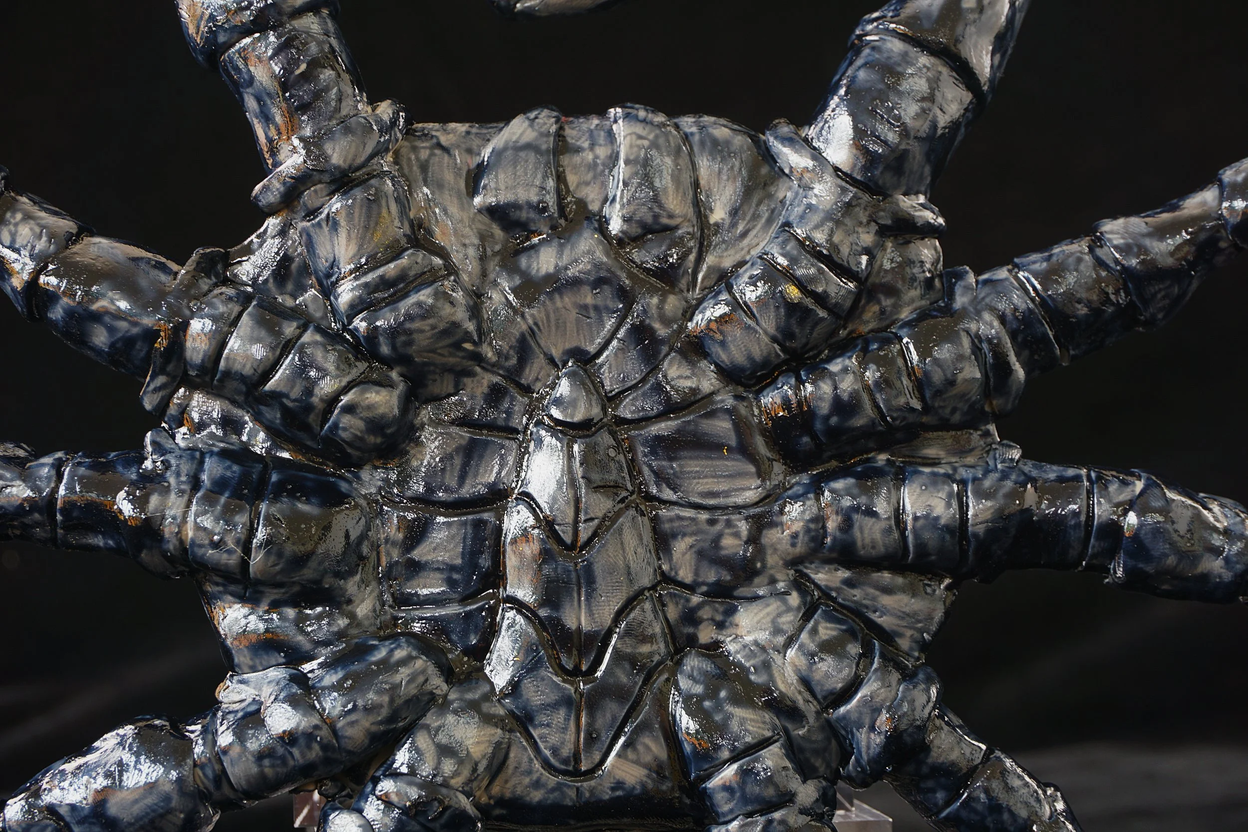 A sculpture of a human face with hands crossing in front, made of reflective metallic material, displayed against a dark background.