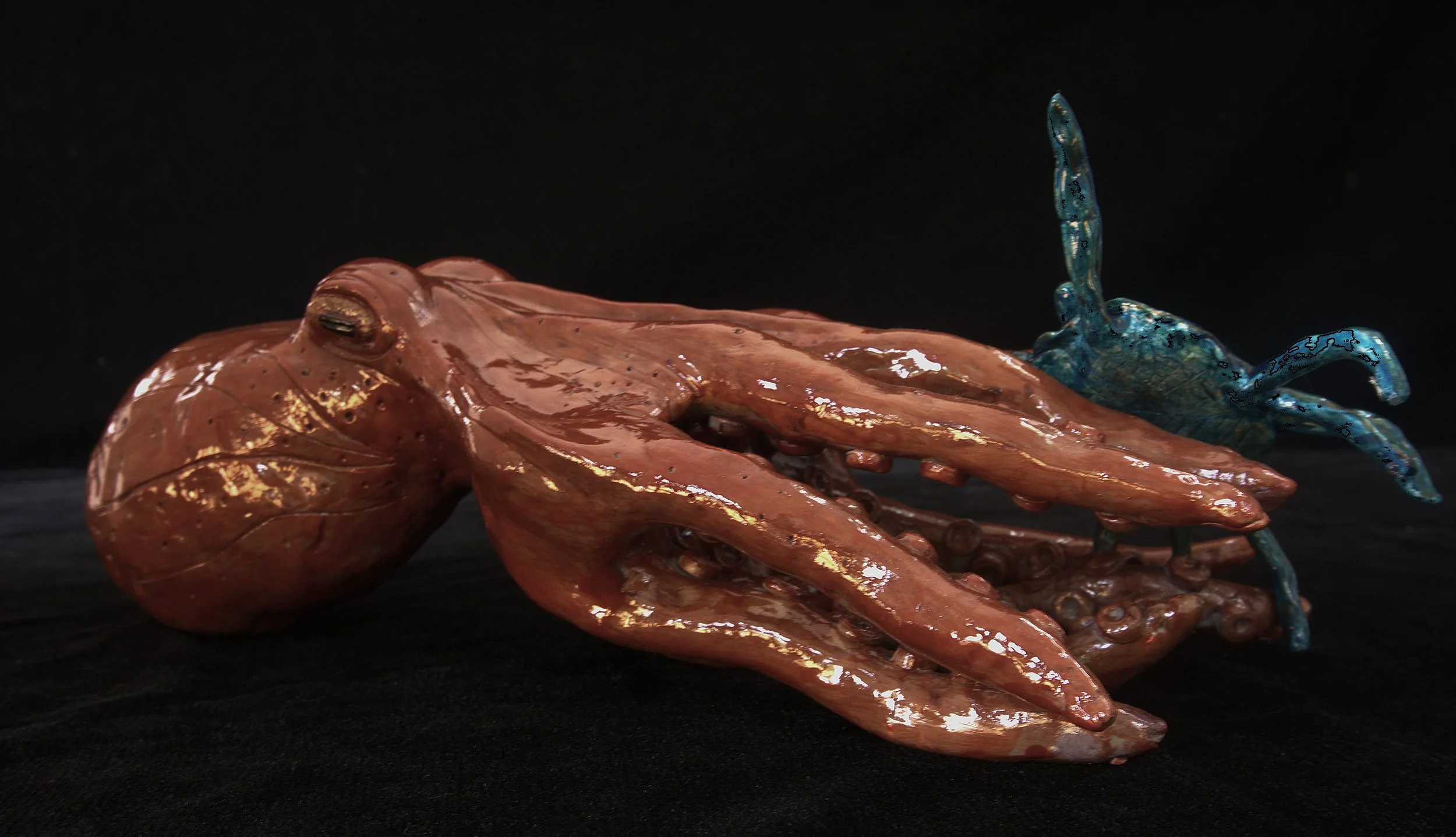 A realistic sculpture of a brown octopus with detailed textures and a blue crab with a shiny blue shell and raised claws, both placed on a black surface against a black background.