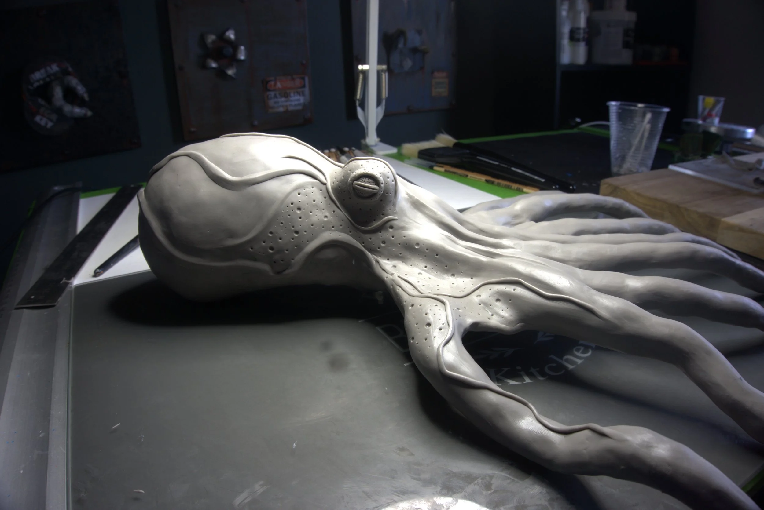 A detailed, realistic sculpture of a squid made from a light-colored material, placed on a work table in a studio or workshop environment with tools and supplies in the background.