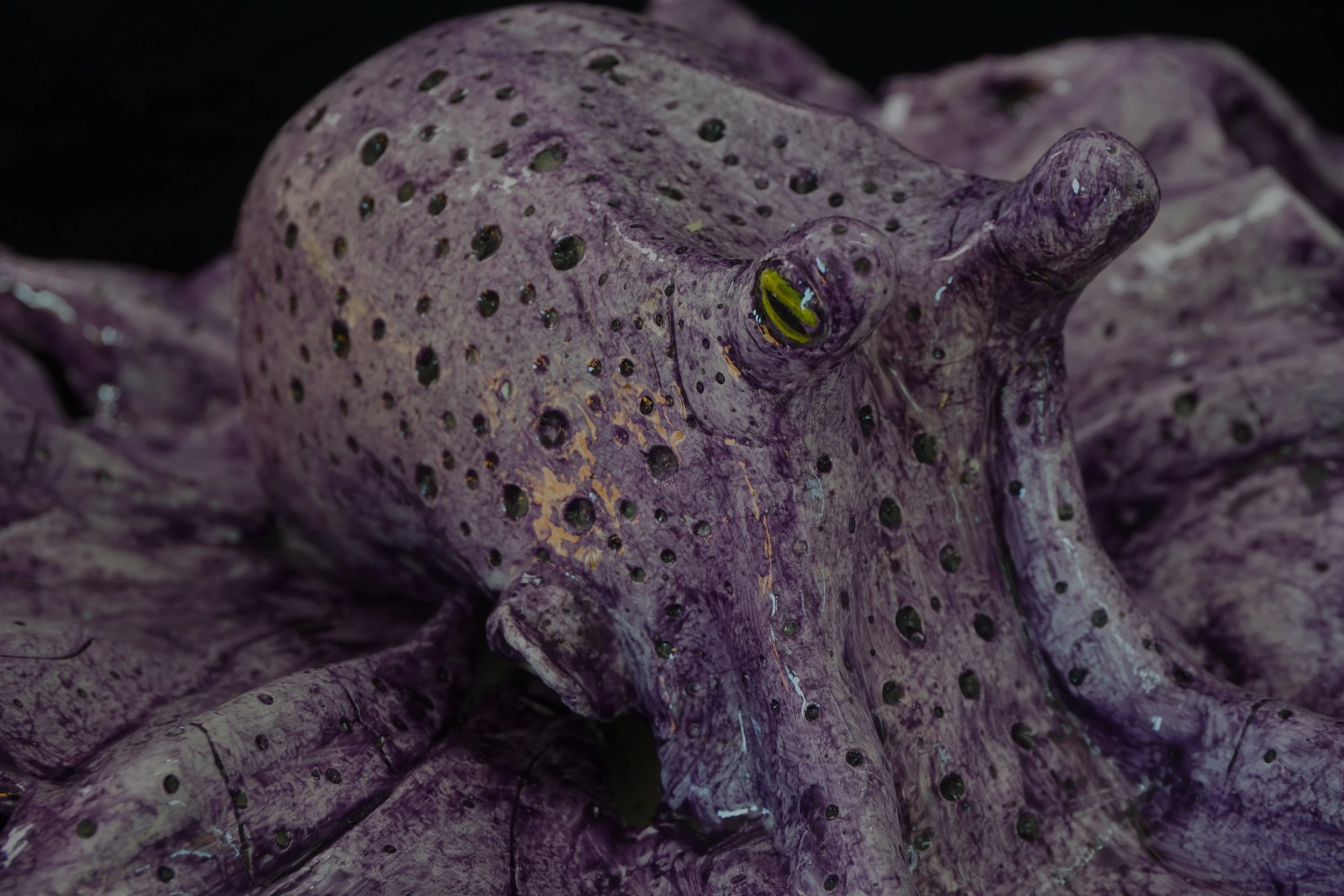 Close-up of a purple ceramic figurine of a chameleon with black spots and greenish-yellow eyes, showing detailed texture.